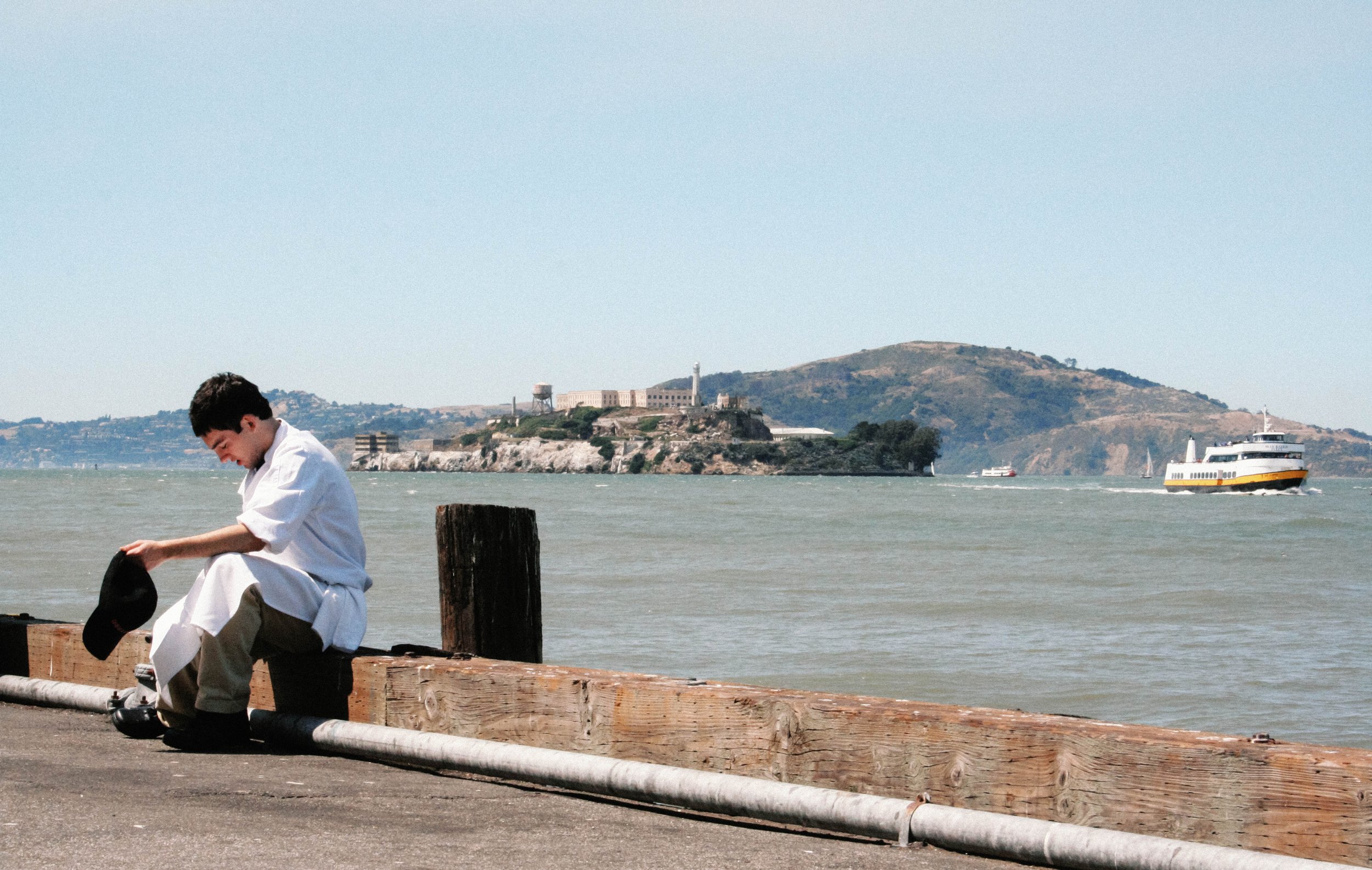 A young boy in a white shirt and khaki pants is sitting on a wooden bench by the water, holding a black hat. In the background, there are boats on the water and a hillside with buildings and a landmark that resembles Alcatraz Island.