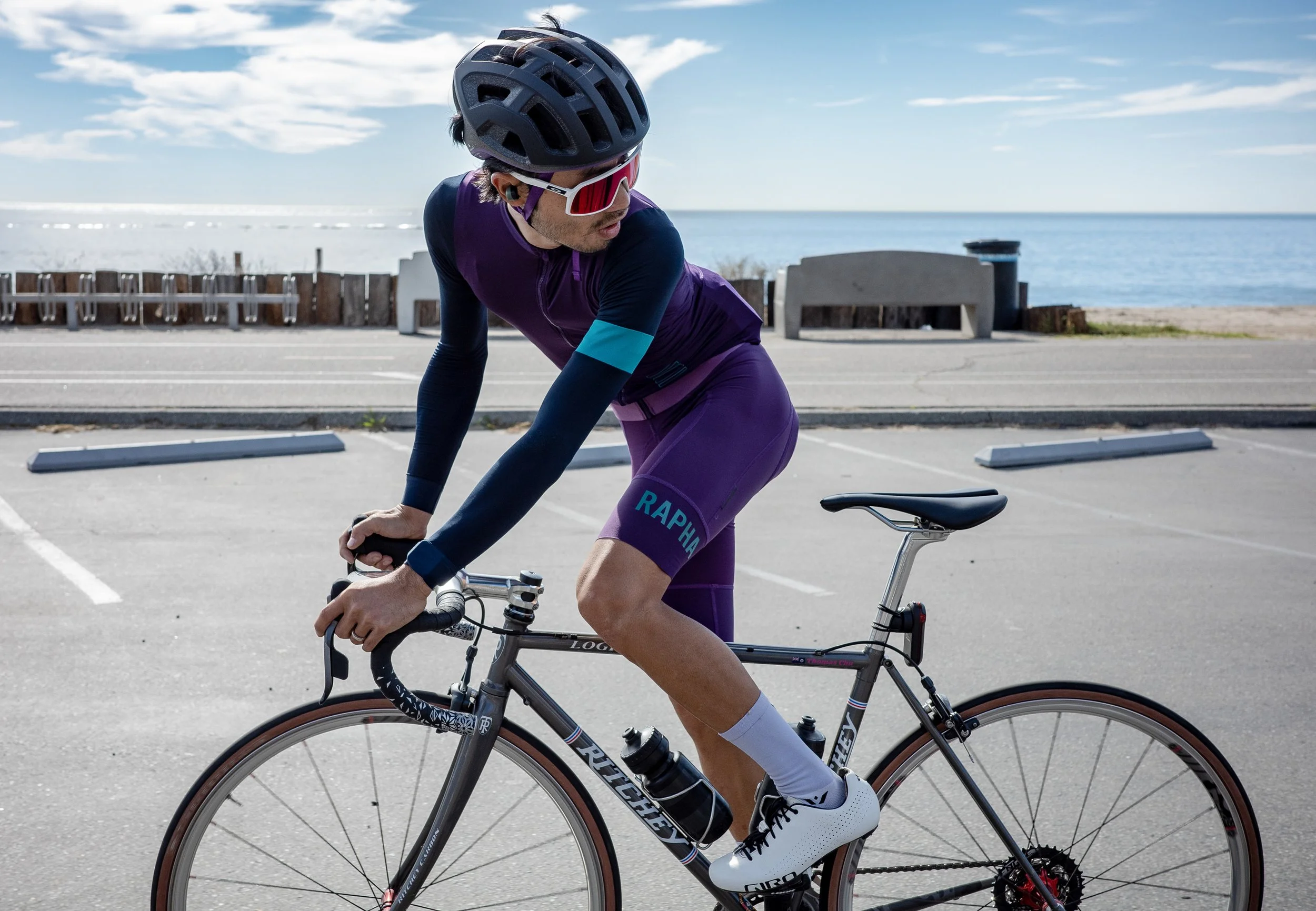 A cyclist wearing a helmet, cycling sunglasses, and a purple cycling outfit, riding a gray bicycle along a paved road with a beach and ocean in the background under a partly cloudy sky.