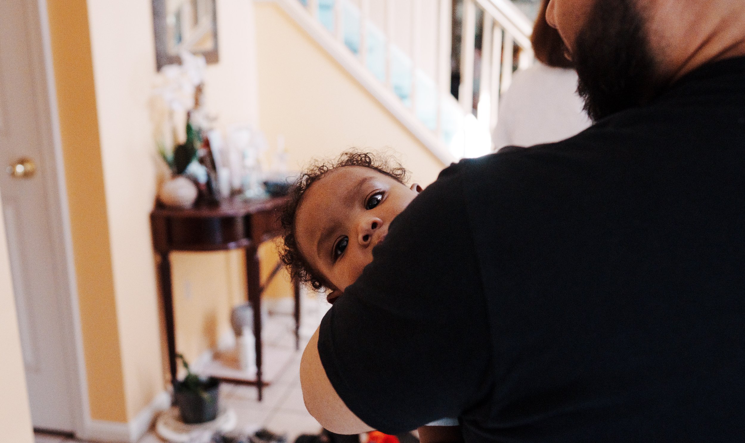 Close-up of a man holding a young child with curly hair, inside a home with yellow walls and a staircase in the background.