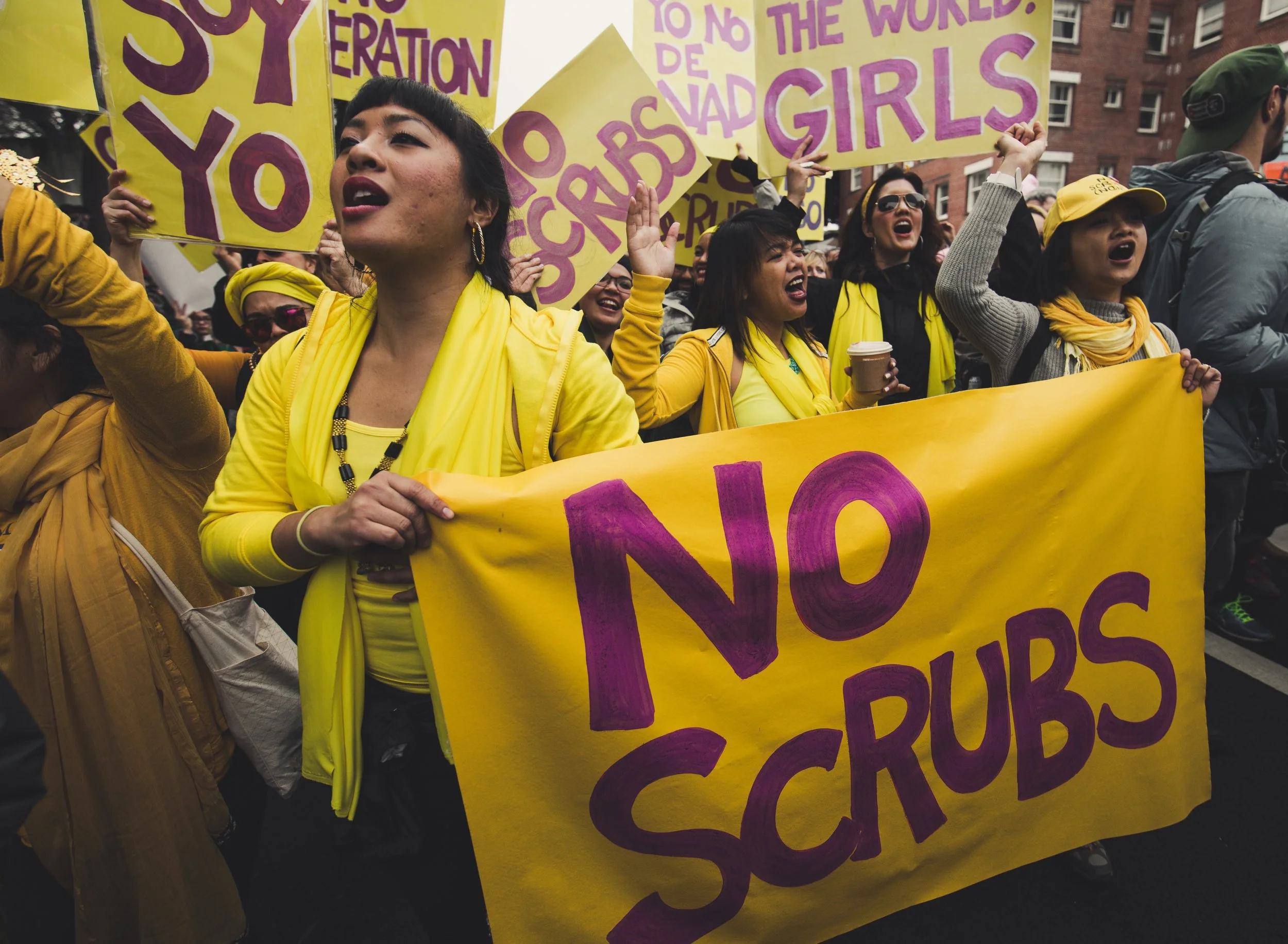 A group of women participating in a protest or march, wearing yellow and holding yellow signs with purple lettering. The signs contain messages advocating for girls' rights and gender equality.