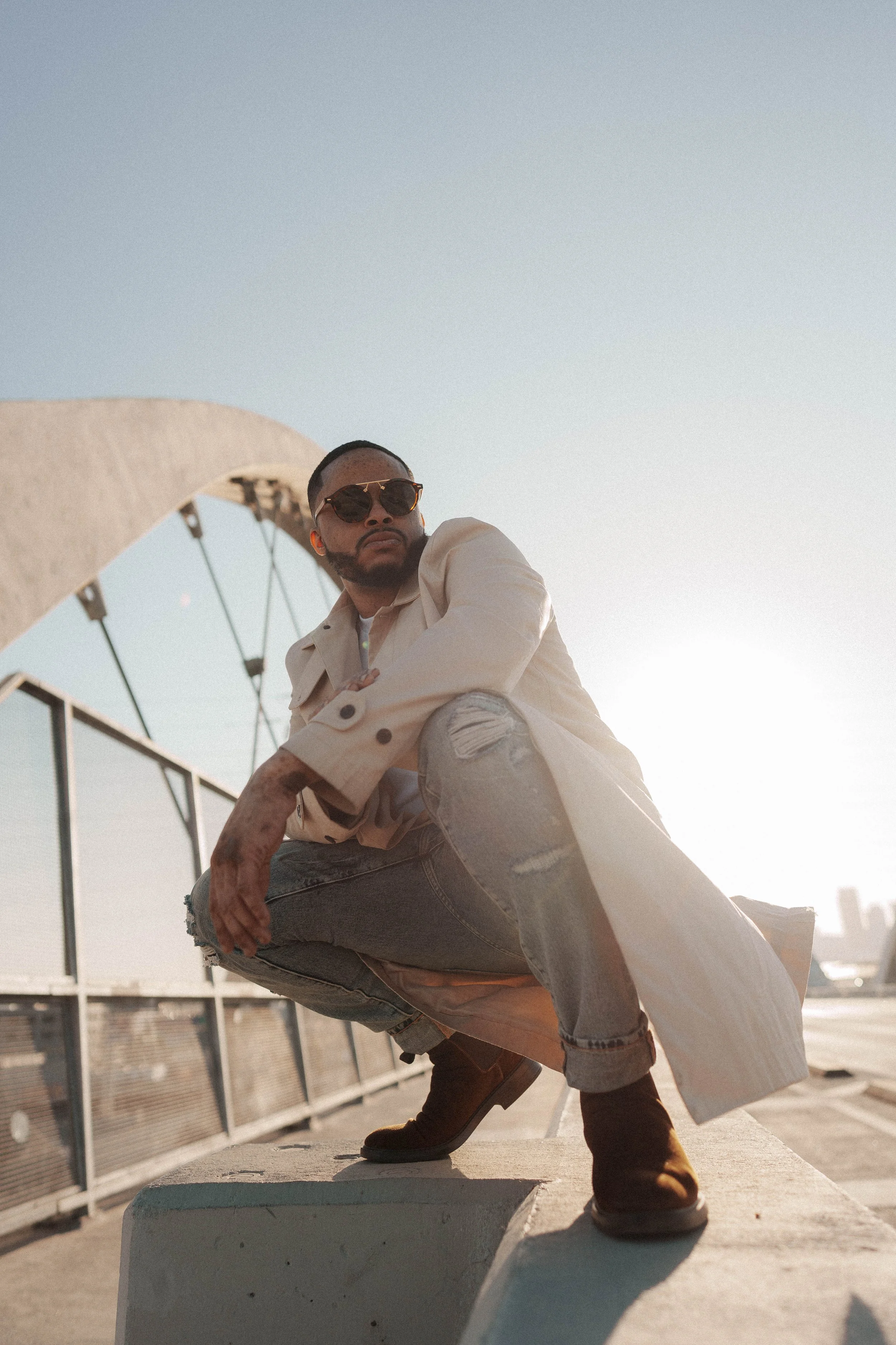 A man crouching on a rooftop with a city skyline in the background, wearing sunglasses, a beige trench coat, ripped jeans, and brown boots.