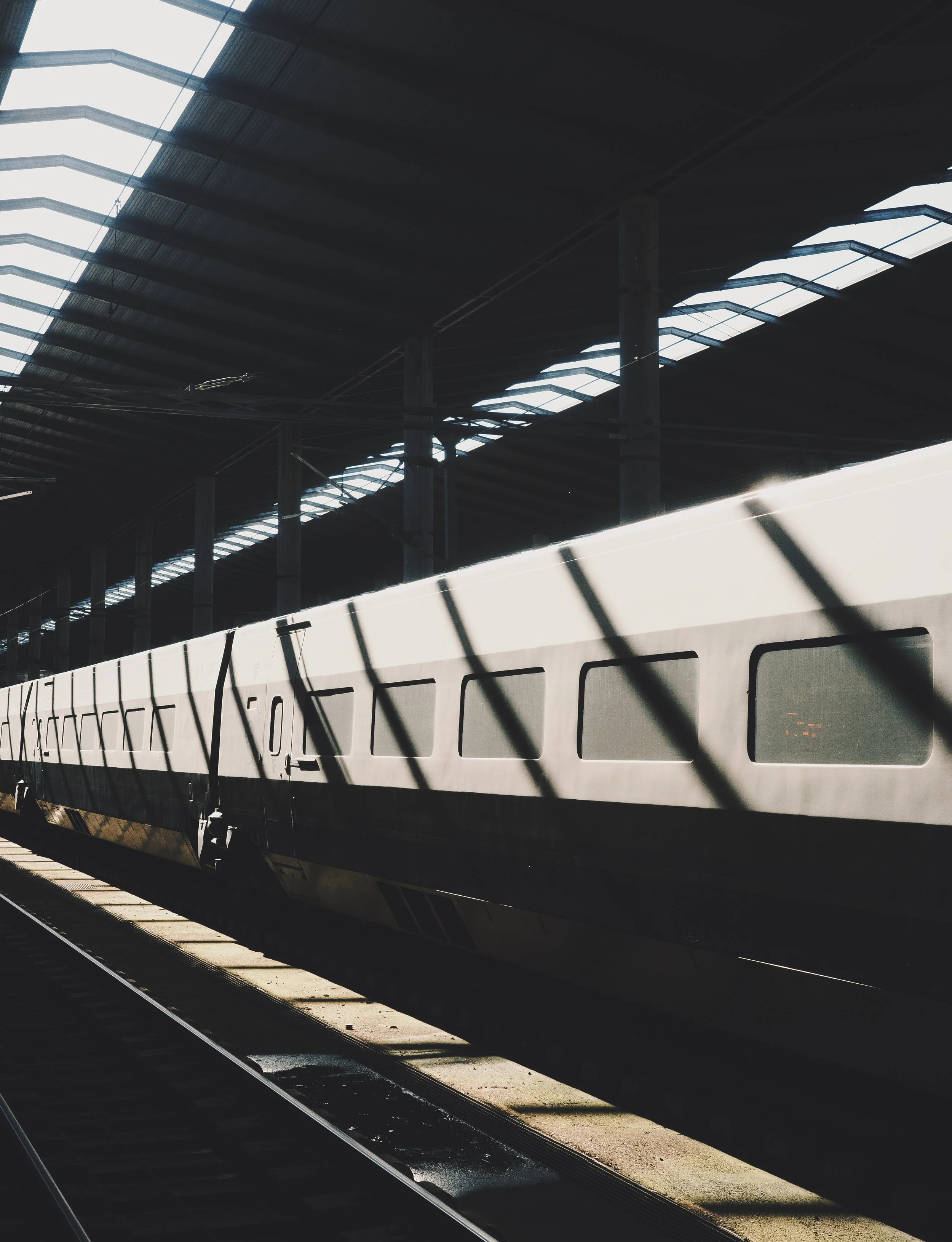 A modern train at a platform with a curved roof and metal supports, casting shadows on the ground and train.