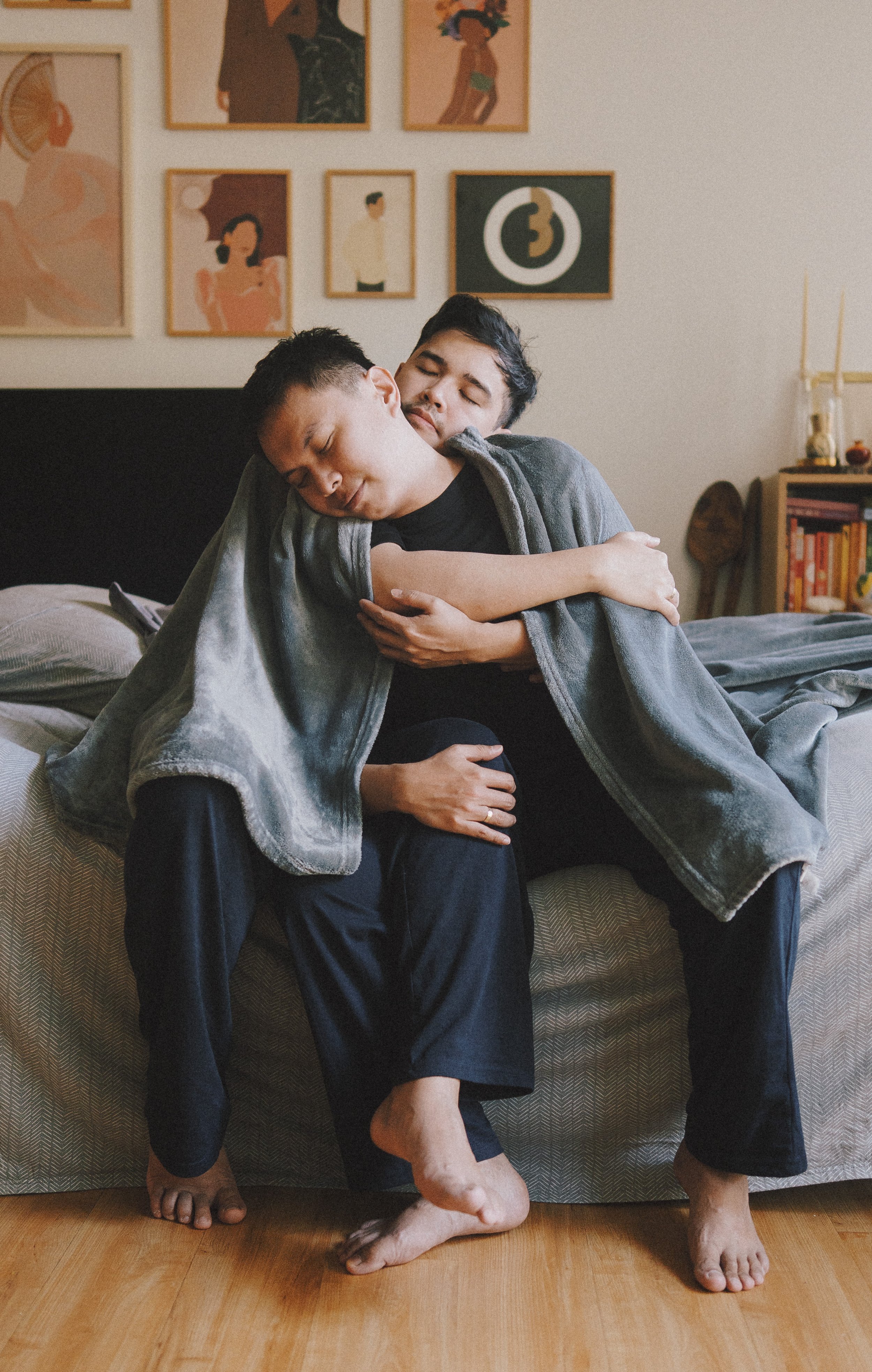 Two men hugging each other on a bed in a cozy room with artwork on the wall and a bookshelf in the background.