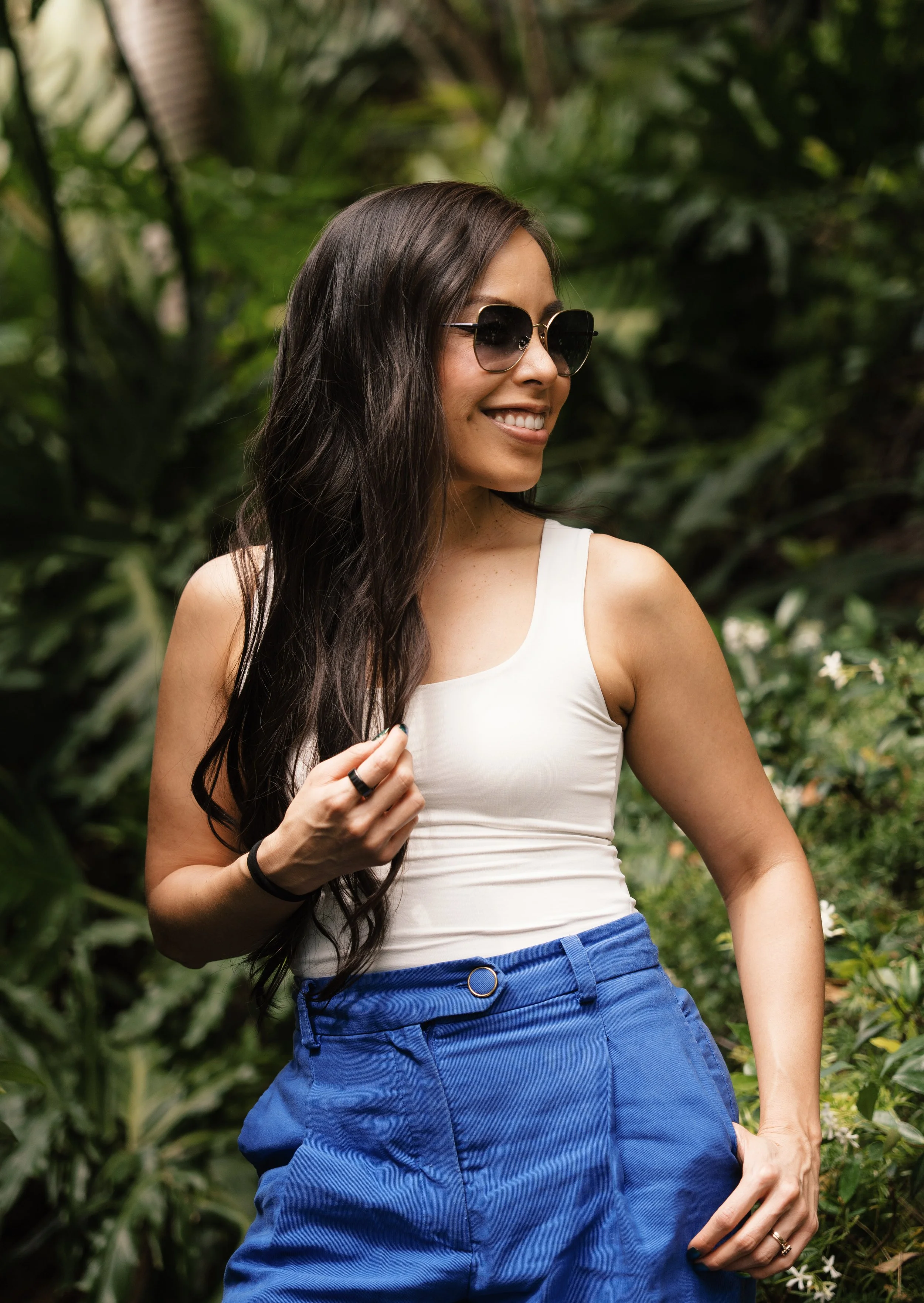 A woman with long dark hair wearing sunglasses, a white sleeveless top, and blue pants, standing outdoors with greenery in the background, smiling and looking to her left.