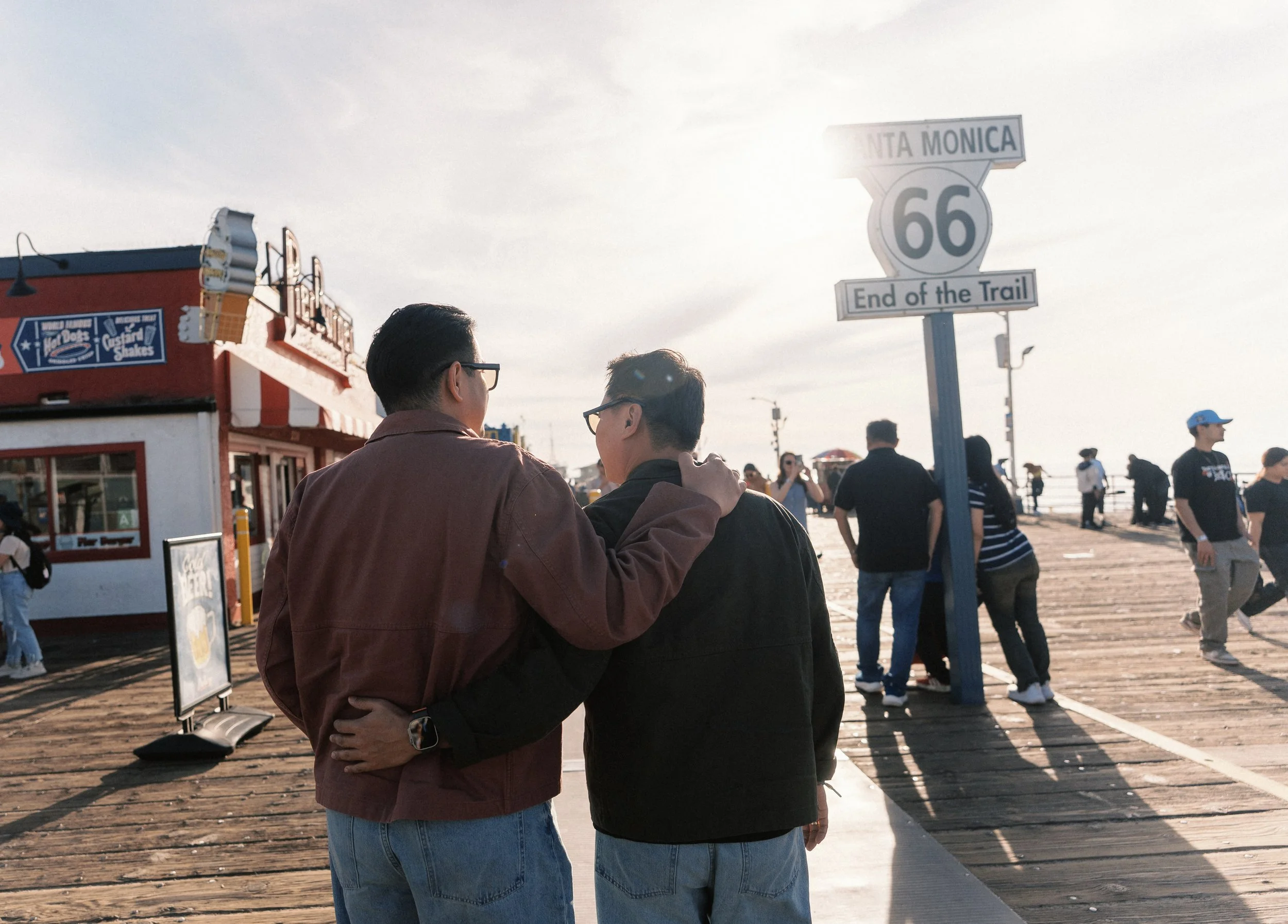 Two young men with glasses and brown and black jackets embrace on a wooden pier at the end of Route 66 in Santa Monica, California, with a sign reading "End of the Trail" and a crowd of people in the background.