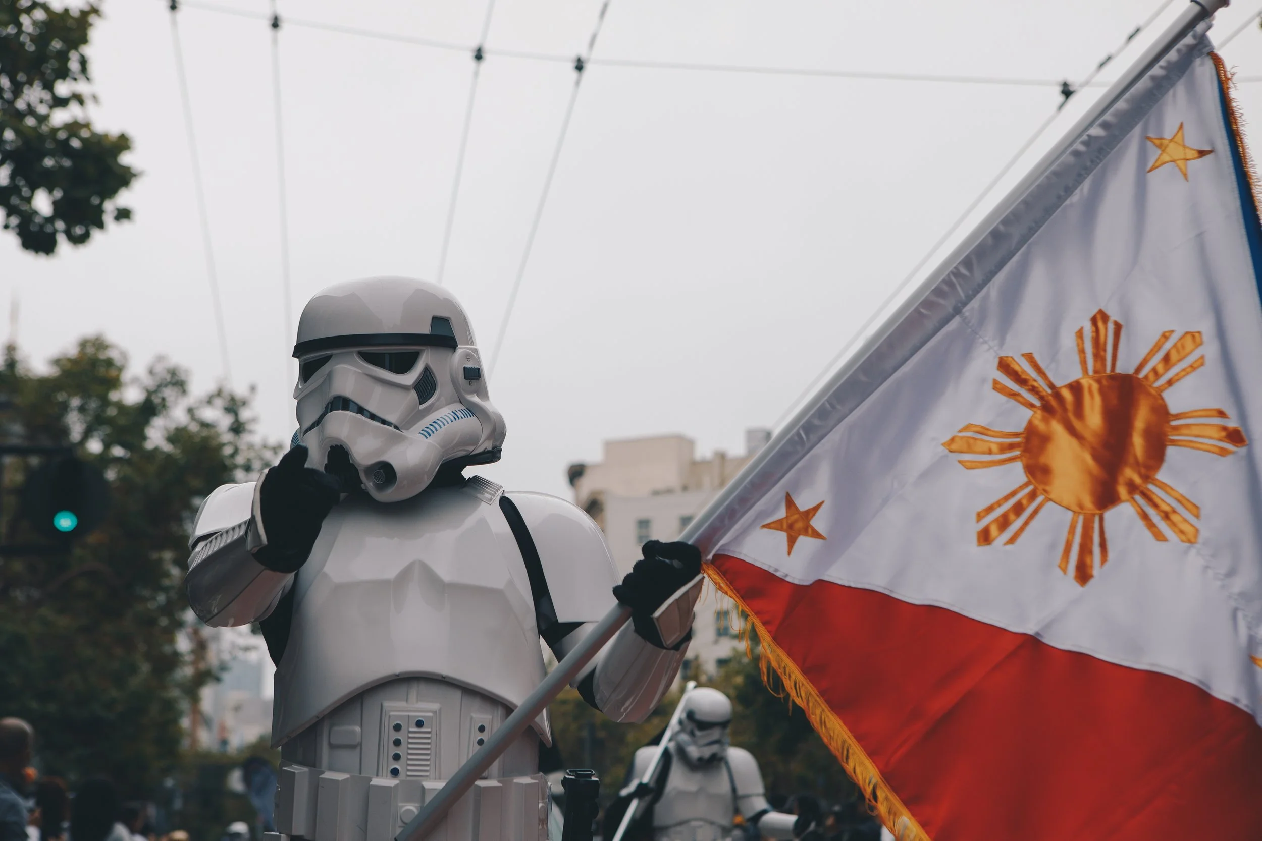 A person dressed as a Star Wars Stormtrooper holding a flag with a golden sun emblem and surrounded by other people during an outdoor event.