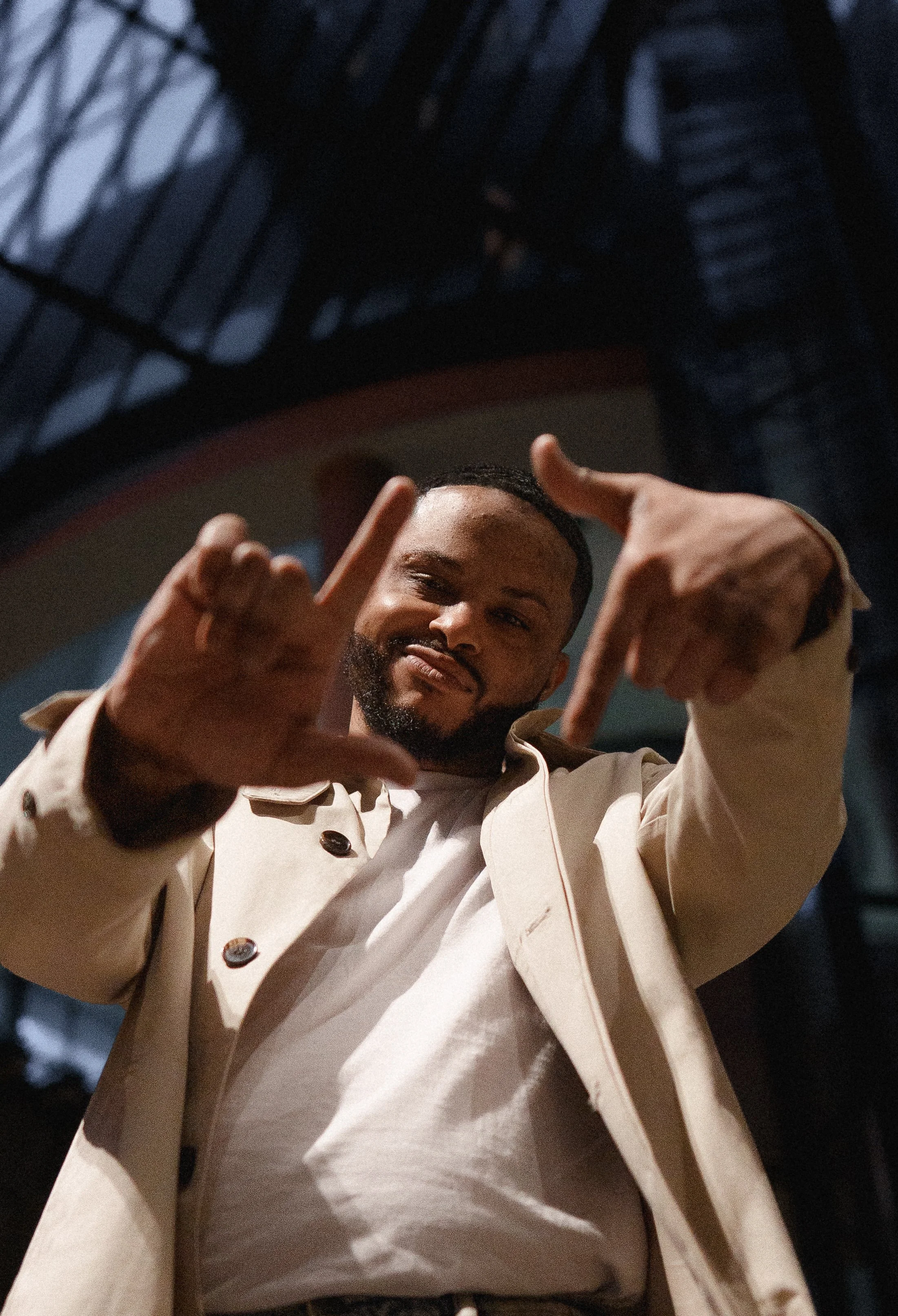 A man with a beard smiling, wearing a beige chef coat, is looking down and making a hand gesture with both hands.
