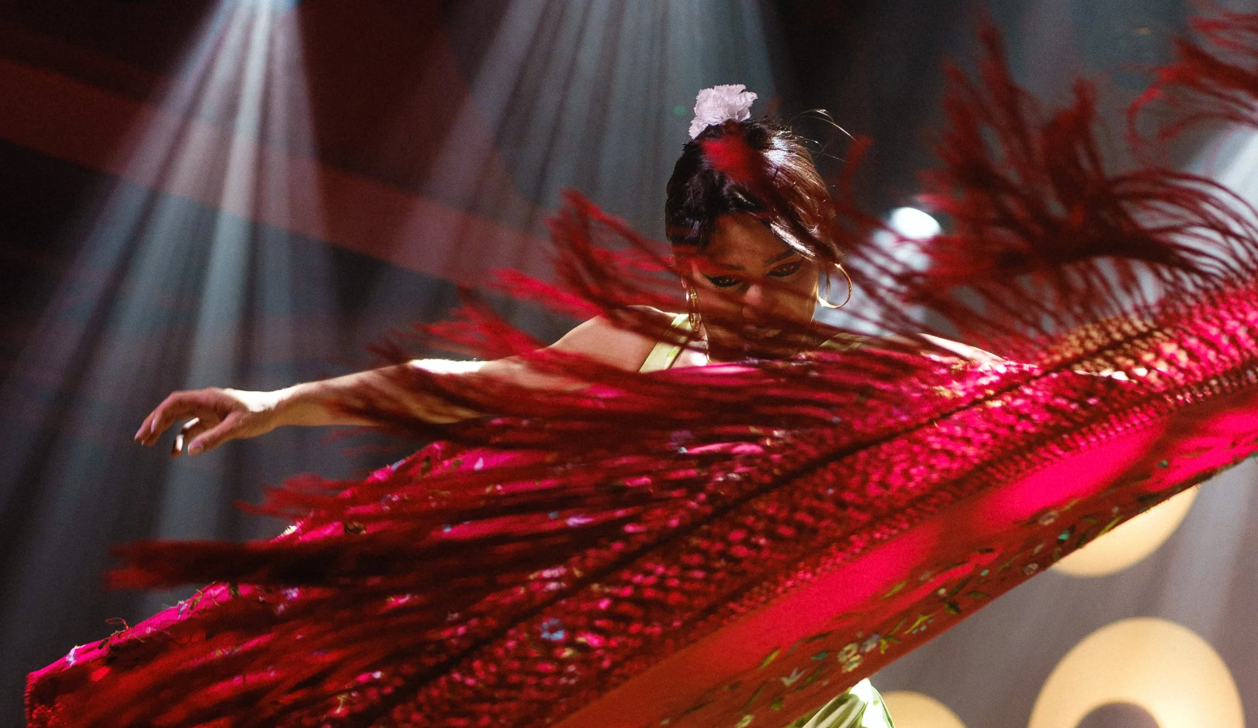 A woman performing a dance on stage, dressed in a vibrant red costume with long fringe, with stage lighting and blurred background.