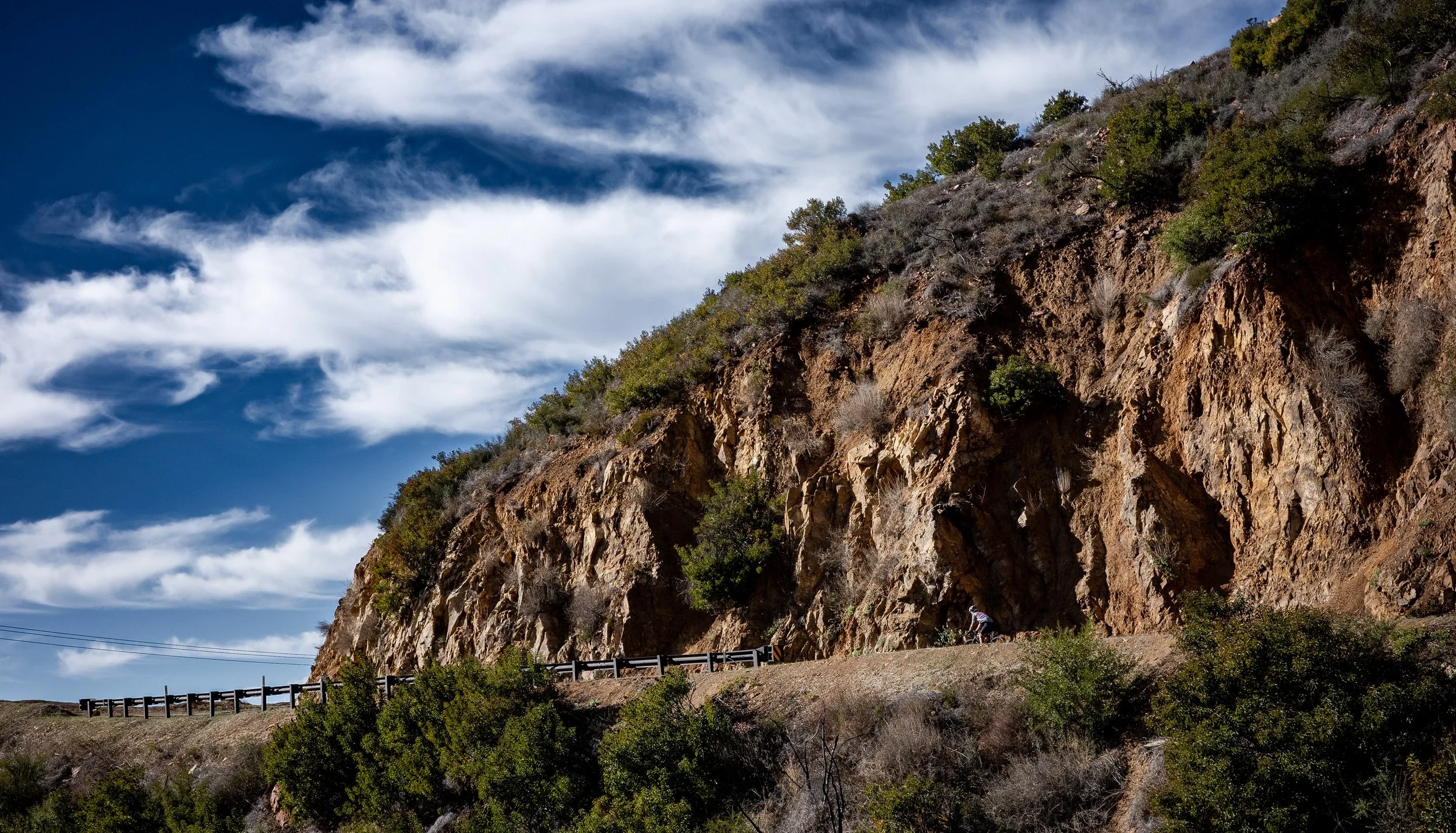 A rocky hillside with sparse green bushes and trees under a blue sky with white clouds. A road with a black guardrail runs along the base of the hill.