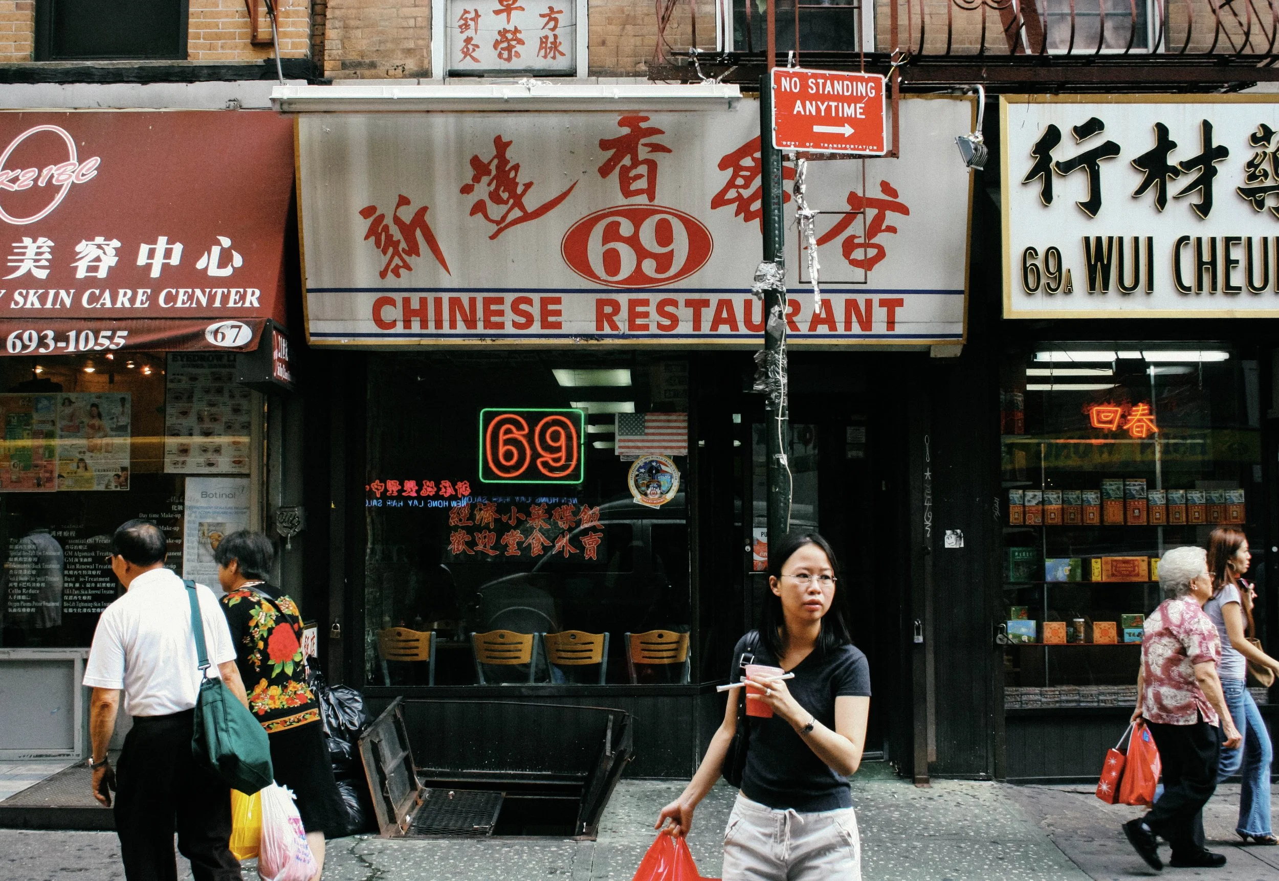 People walking on a city street in front of Chinese restaurant signs, including one with red characters and the number 69, a skin care center sign in Chinese and English, and a WU CHEUNG sign with Chinese characters and the address 69A.