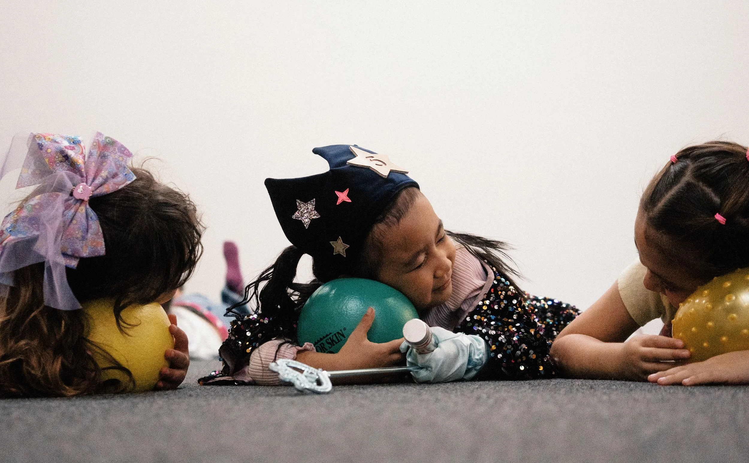 A group of children sitting on the floor, holding balloons, with one girl wearing a unicorn-themed party hat and a girl with a large colorful bow in her hair, engaged in a celebration.