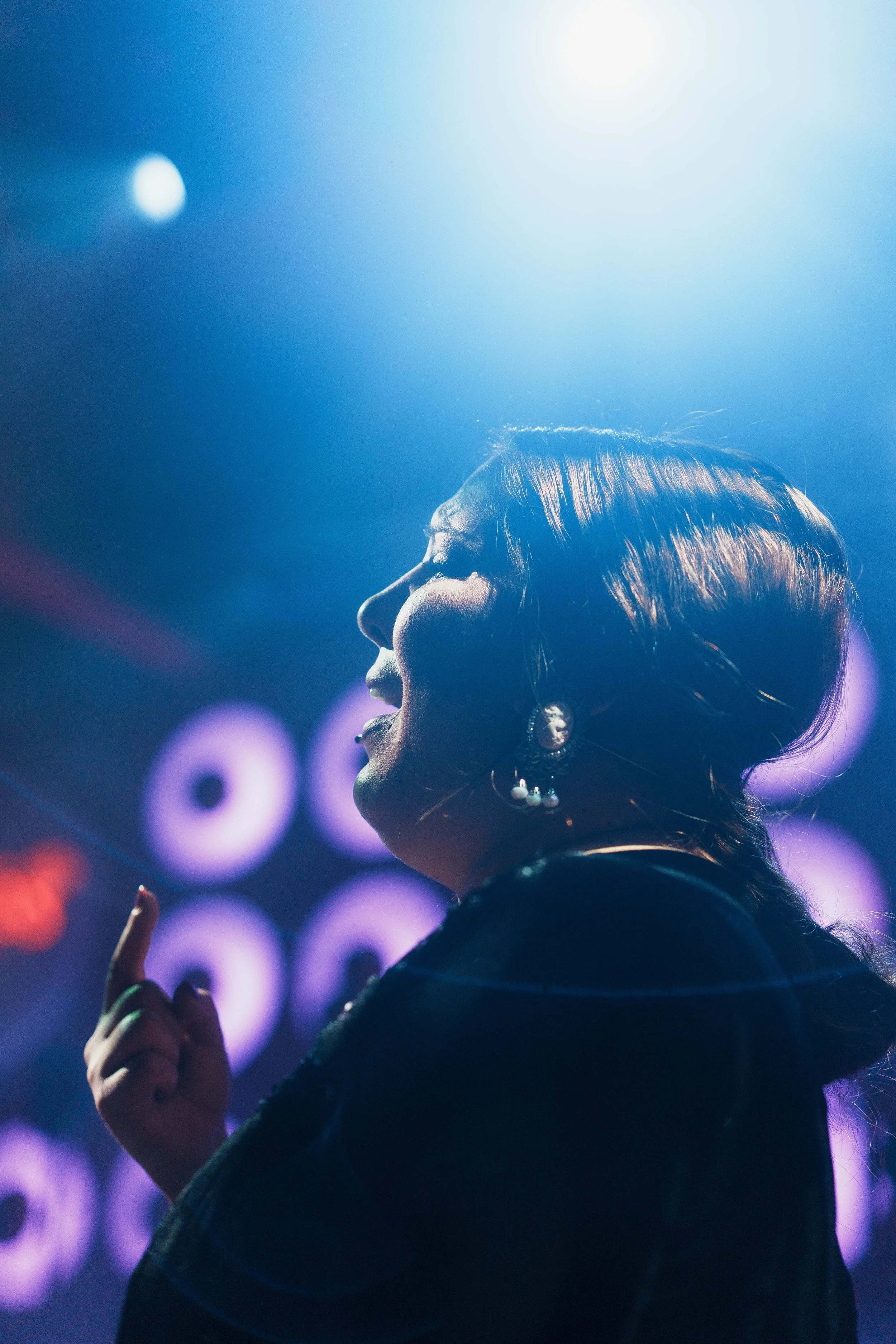 A woman with brown hair styled in soft waves, wearing earrings, is captured in profile with her eyes closed, smiling, and appearing joyful while facing bright lights in the background at a vibrant event.