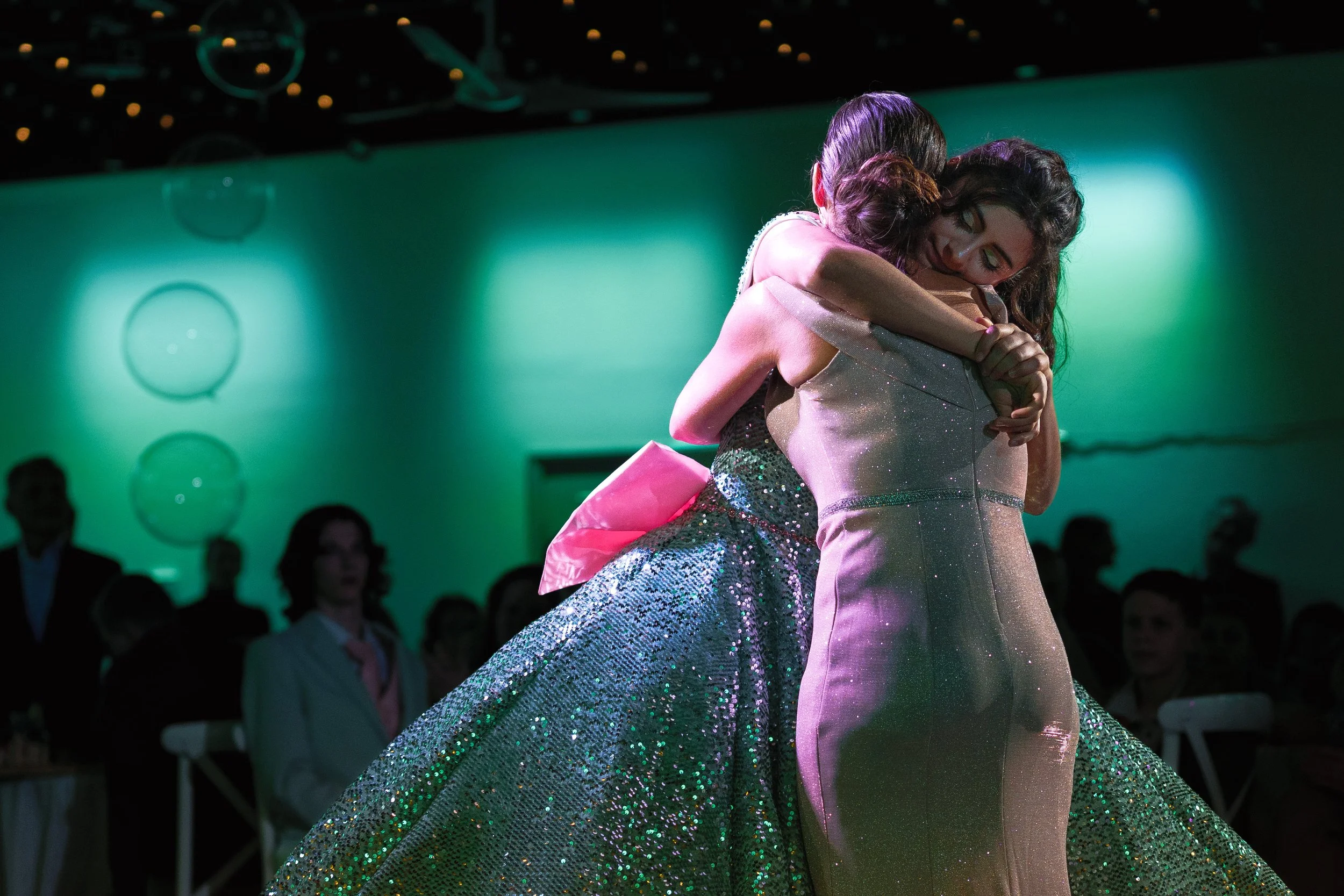 Two women hugging each other on a stage with green lighting, wearing glamorous dresses, in front of an audience.