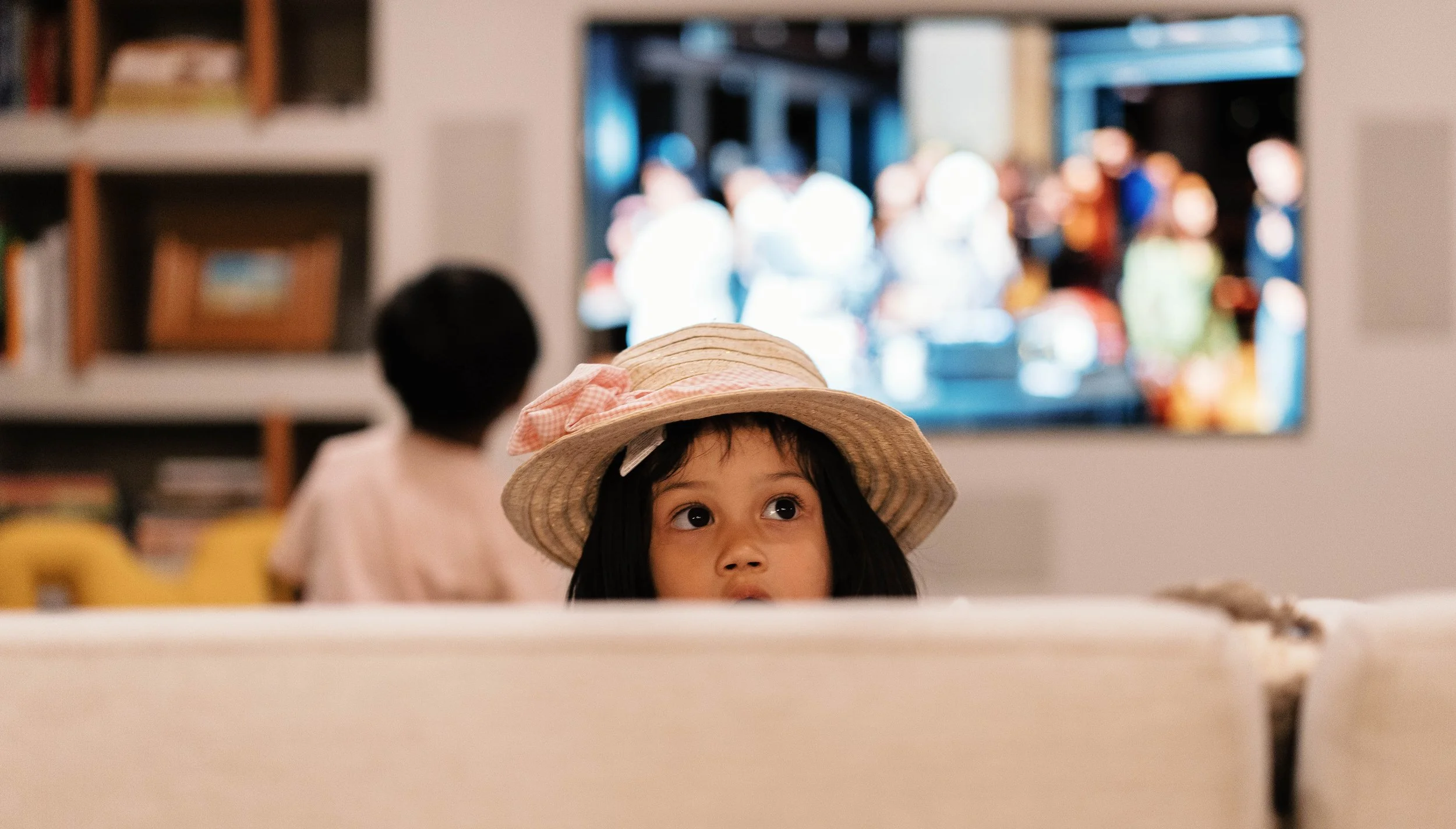Young girl wearing a straw hat looking upward with a monitor showing a crowded scene in the background.