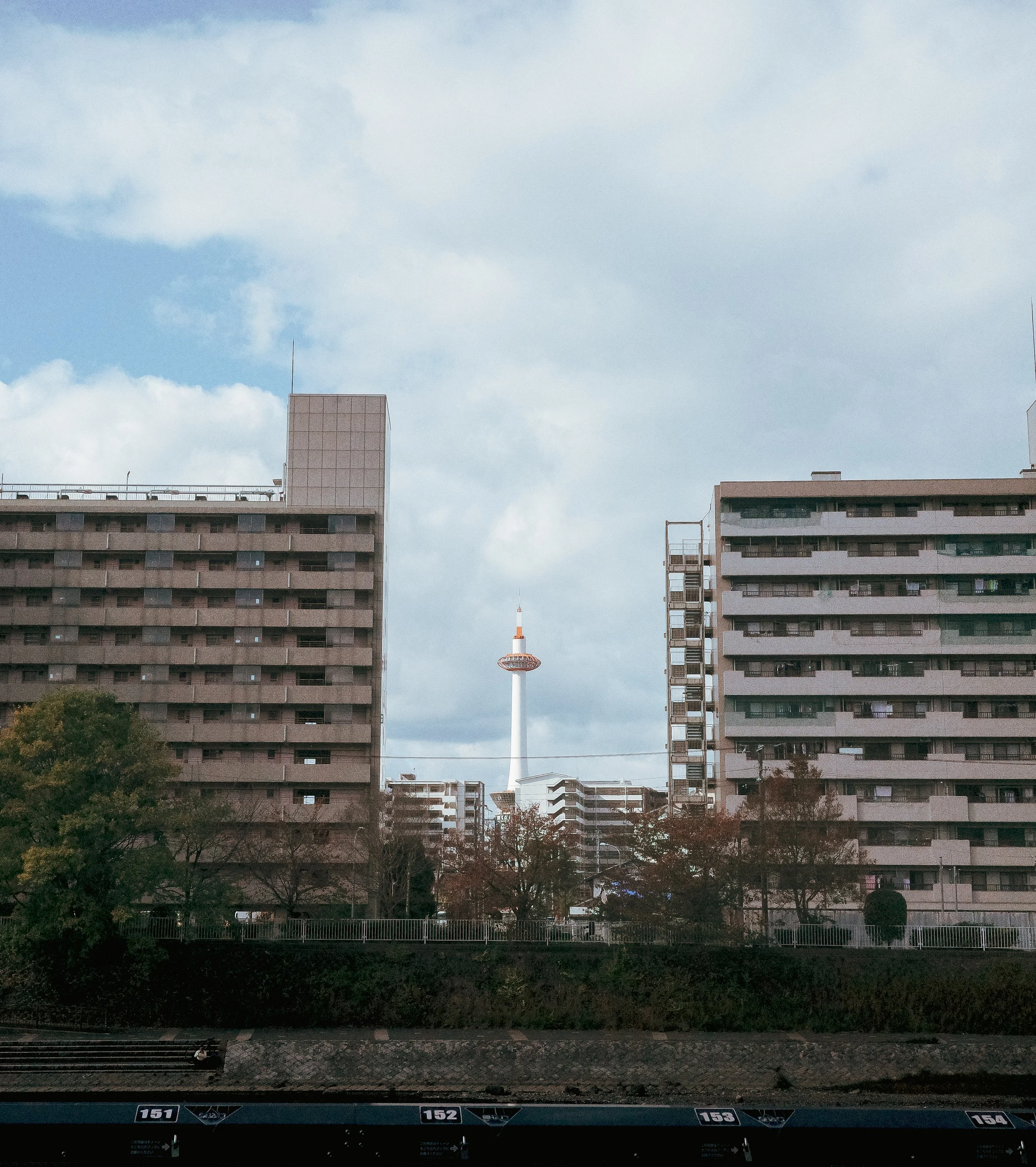 Cityscape view with apartment buildings, trees, and a telecommunications tower in the background under a mostly cloudy sky.