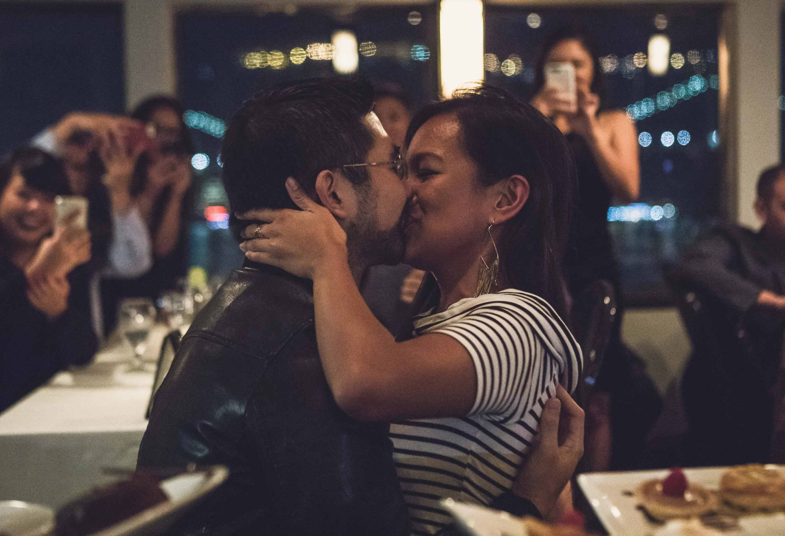 A couple kissing at a celebration or party with guests taking photos in the background.