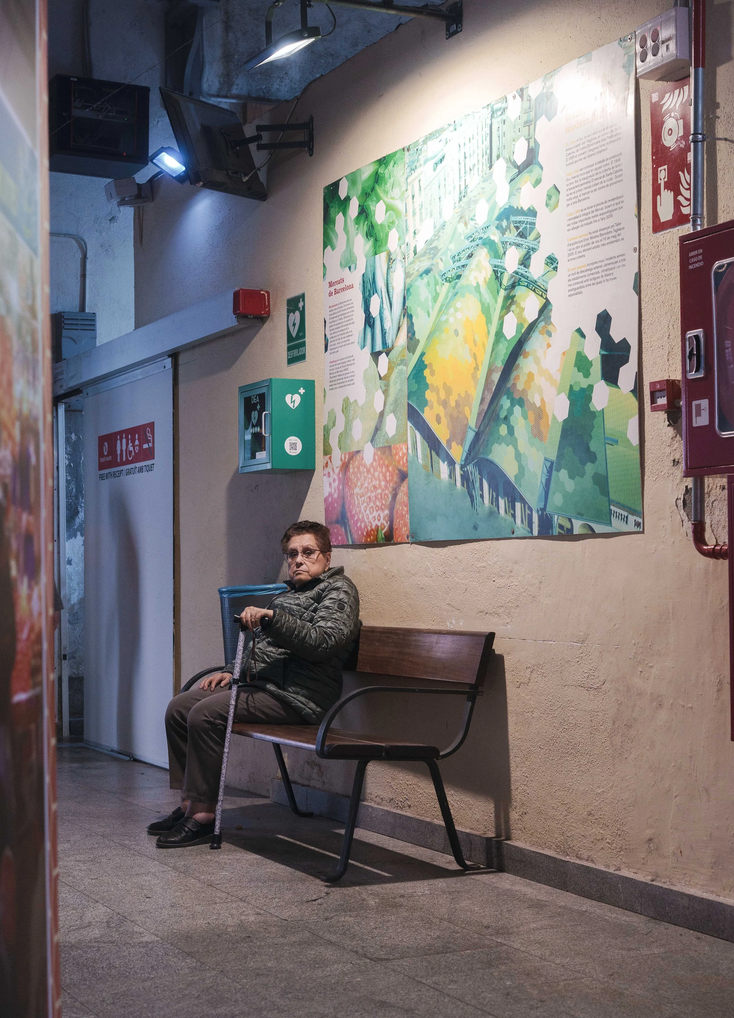 An elderly woman wearing glasses and a black jacket sits on a wooden bench, holding a cane, in a waiting area. Behind her is a colorful mural on the wall, featuring abstract foliage and architectural elements. The corridor has tiled flooring, fire sa