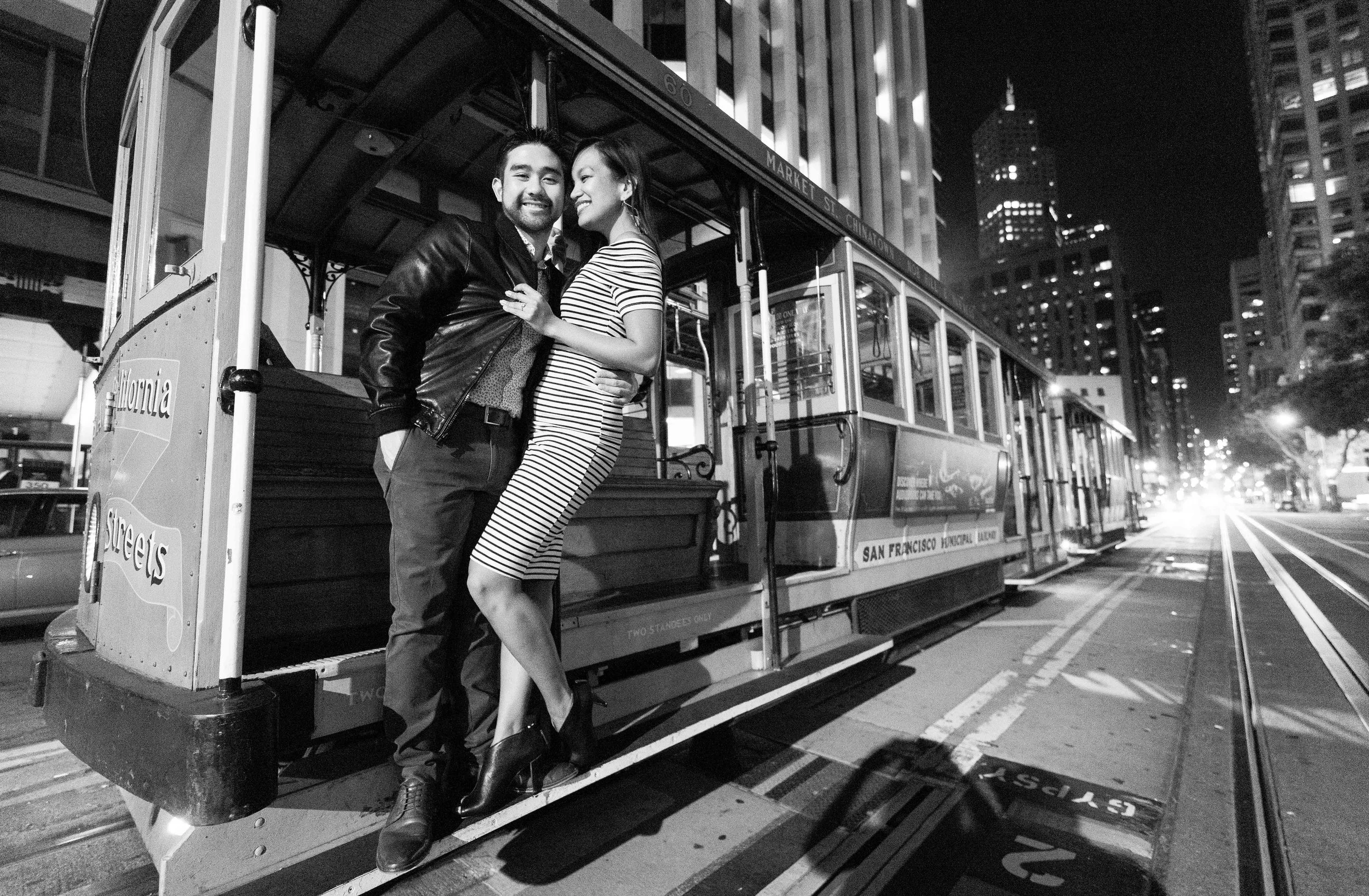 A couple smiling and hugging on a San Francisco cable car at night, with tall buildings in the background.