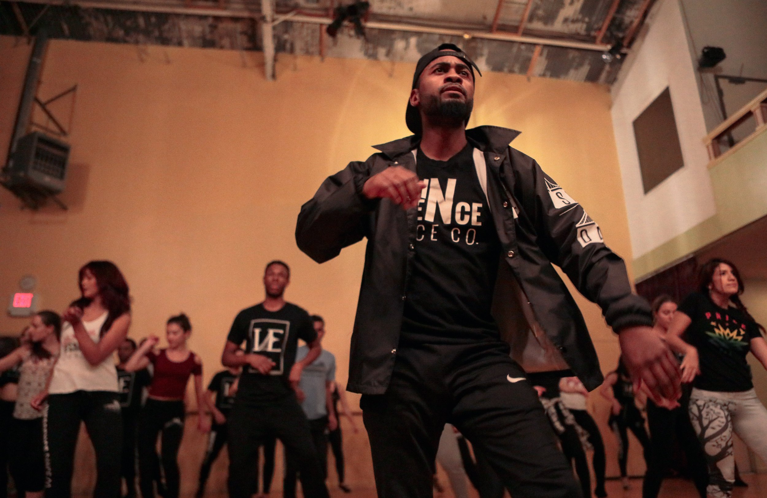 A man wearing a black jacket, black cap backwards, and black t-shirt with white text is dancing among a group of people in a room with beige walls and a high ceiling, with other people standing and sitting around him.
