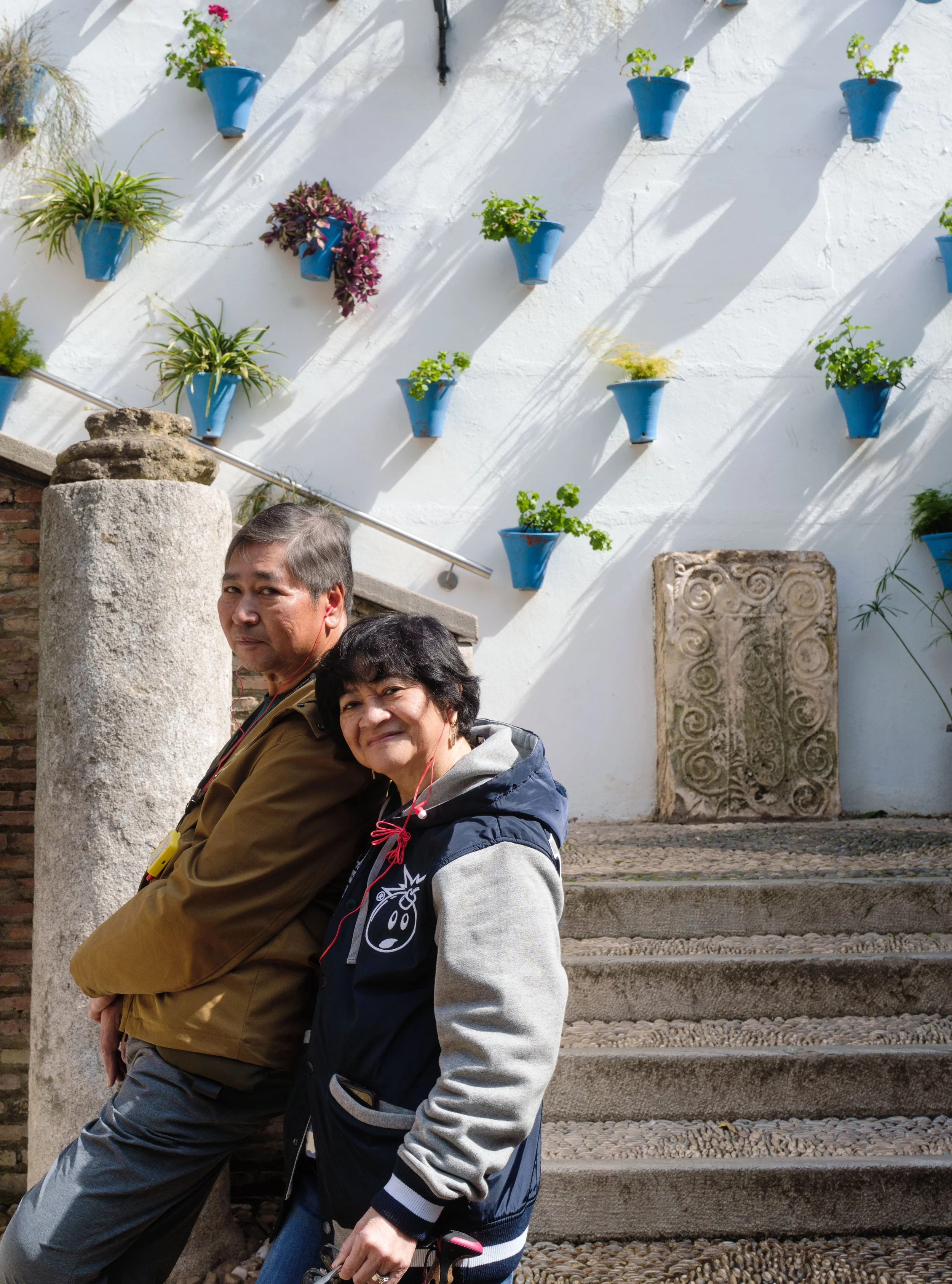 Two people sitting on stone stairs in front of a white wall decorated with small blue pots of flowering plants.