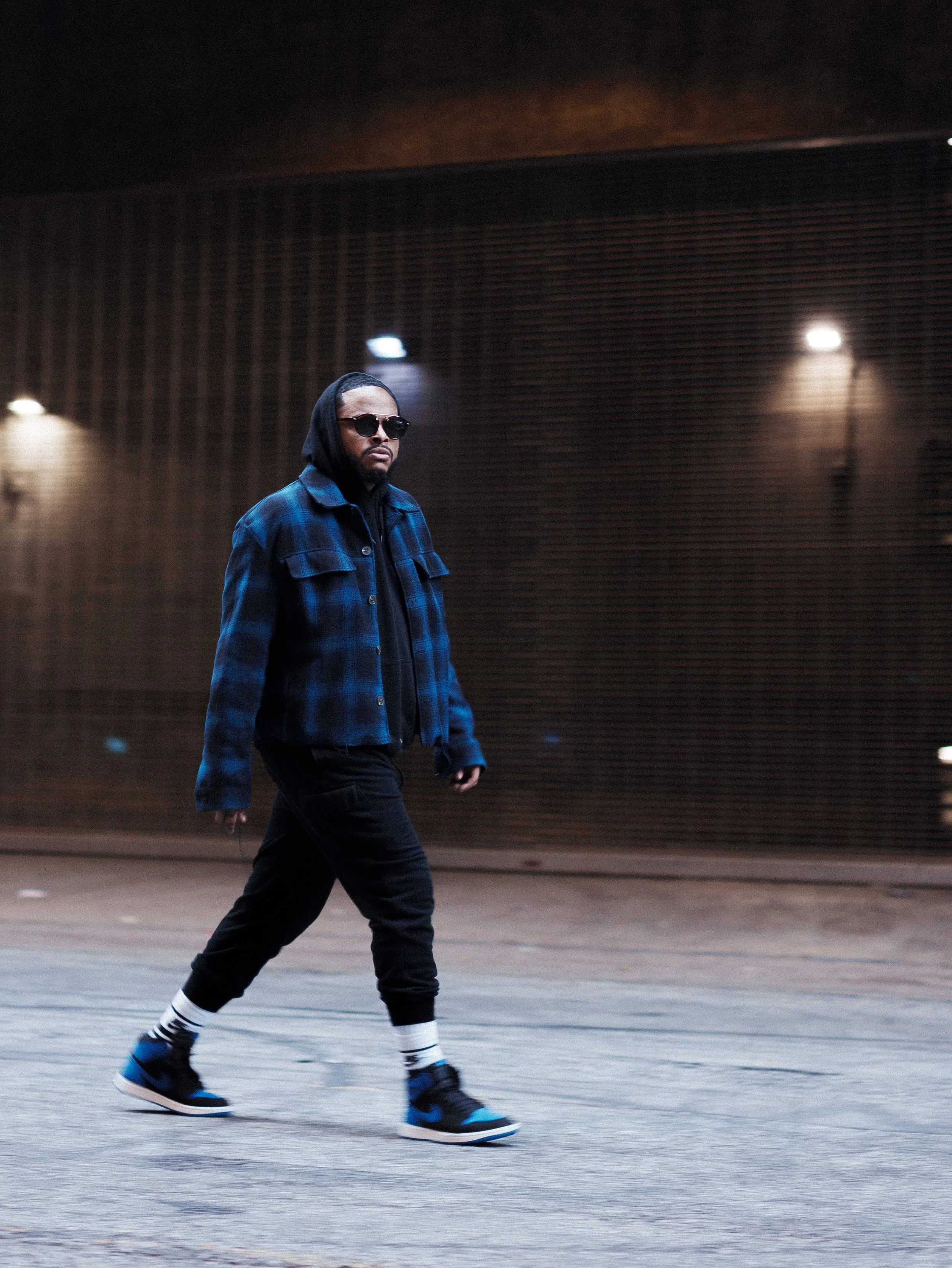 A man walking across an urban street at night, wearing sunglasses, a blue plaid jacket, black pants, and sneakers, with a dark background and streetlights lighting the area.