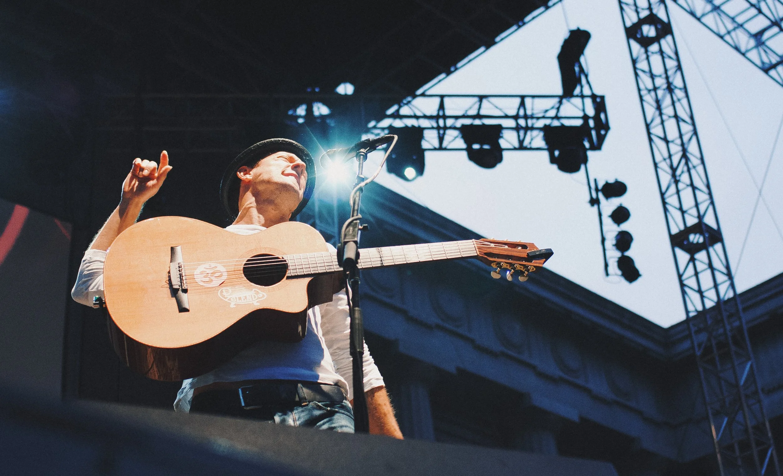 Jason Mraz playing guitar on stage during outdoor concert with stage lights and equipment overhead.