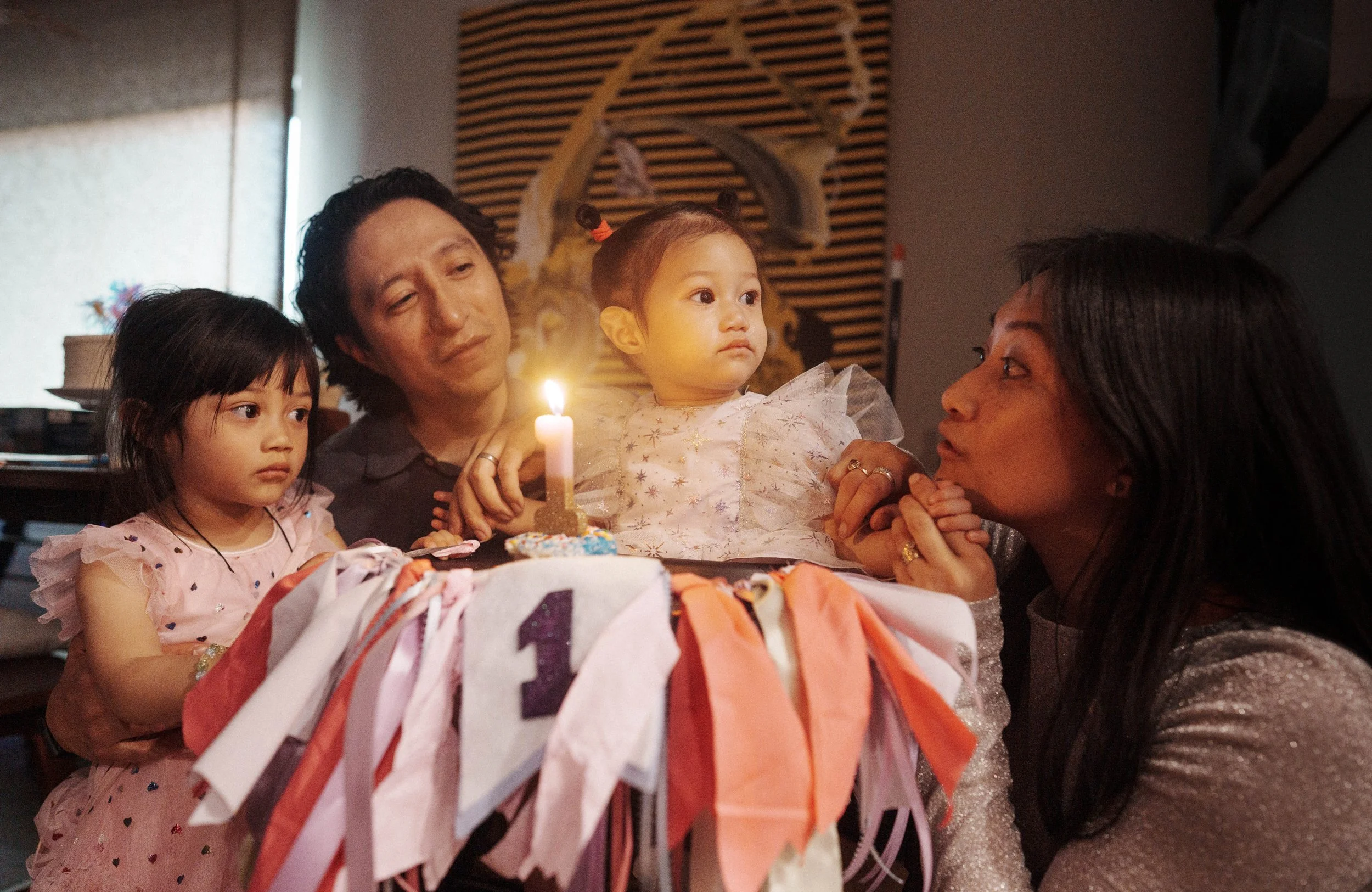 A group of five people gathered around a birthday cake with a lit candle, celebrating a child's birthday. The group includes two young girls, a man, and two women. The setting appears to be indoors with a decorative wooden wall in the background.