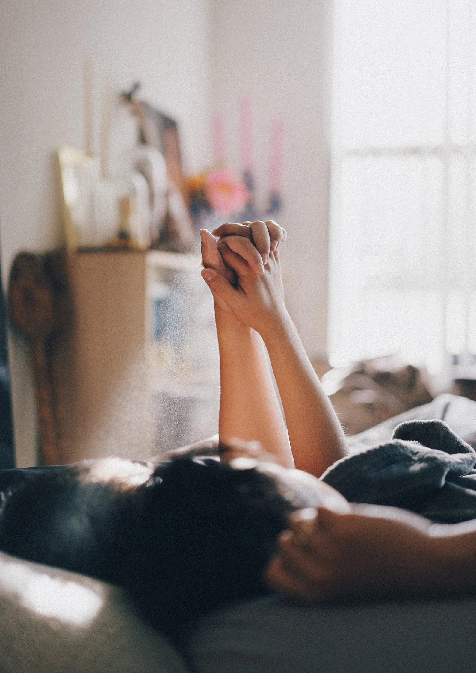 A person with hands clasped together resting on their chest, lying in bed with a blurred background of a room with a dresser and a window.