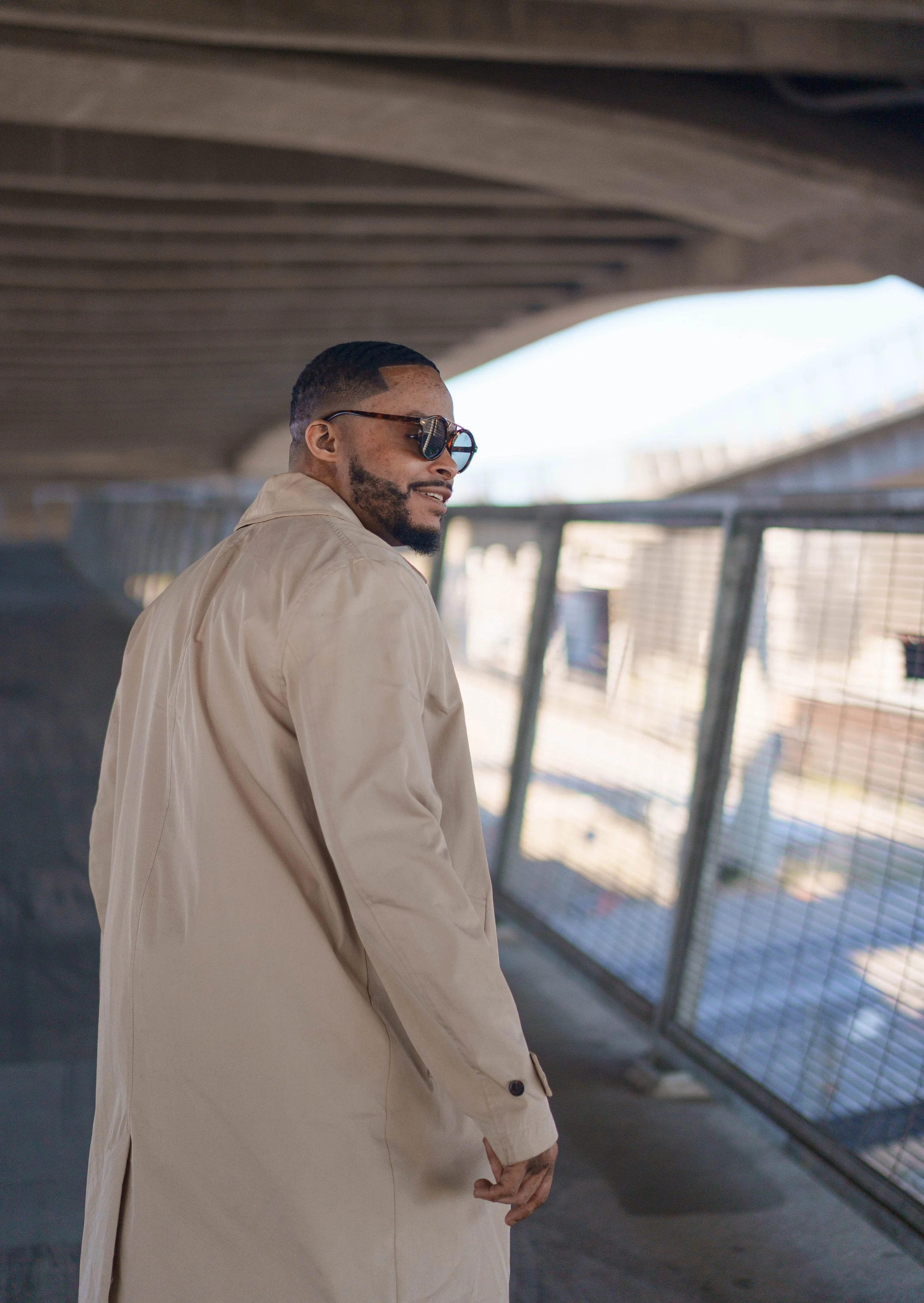 A man in a beige trench coat and sunglasses standing in an urban setting under a bridge or overpass, with blurred cityscape and fence in the background.