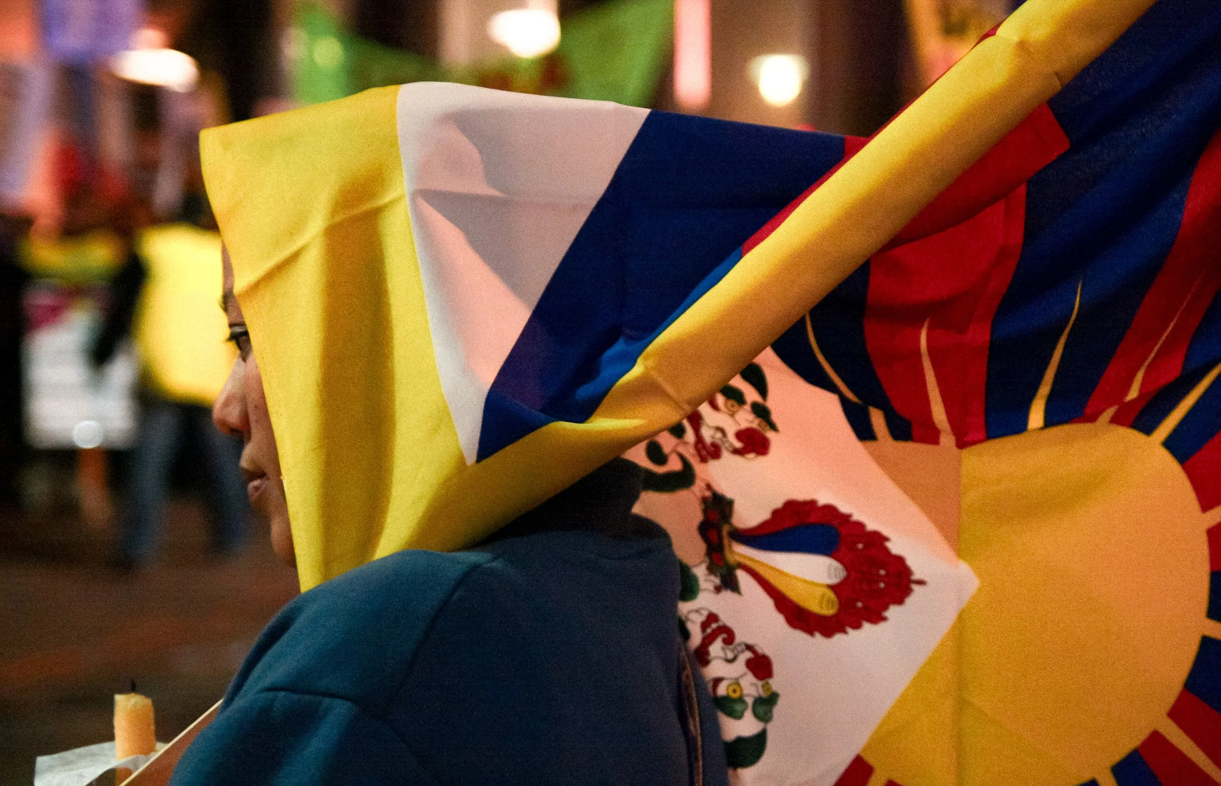 Person holding a Mexican flag during a celebration or event, with a blurred background of colorful lights and people.