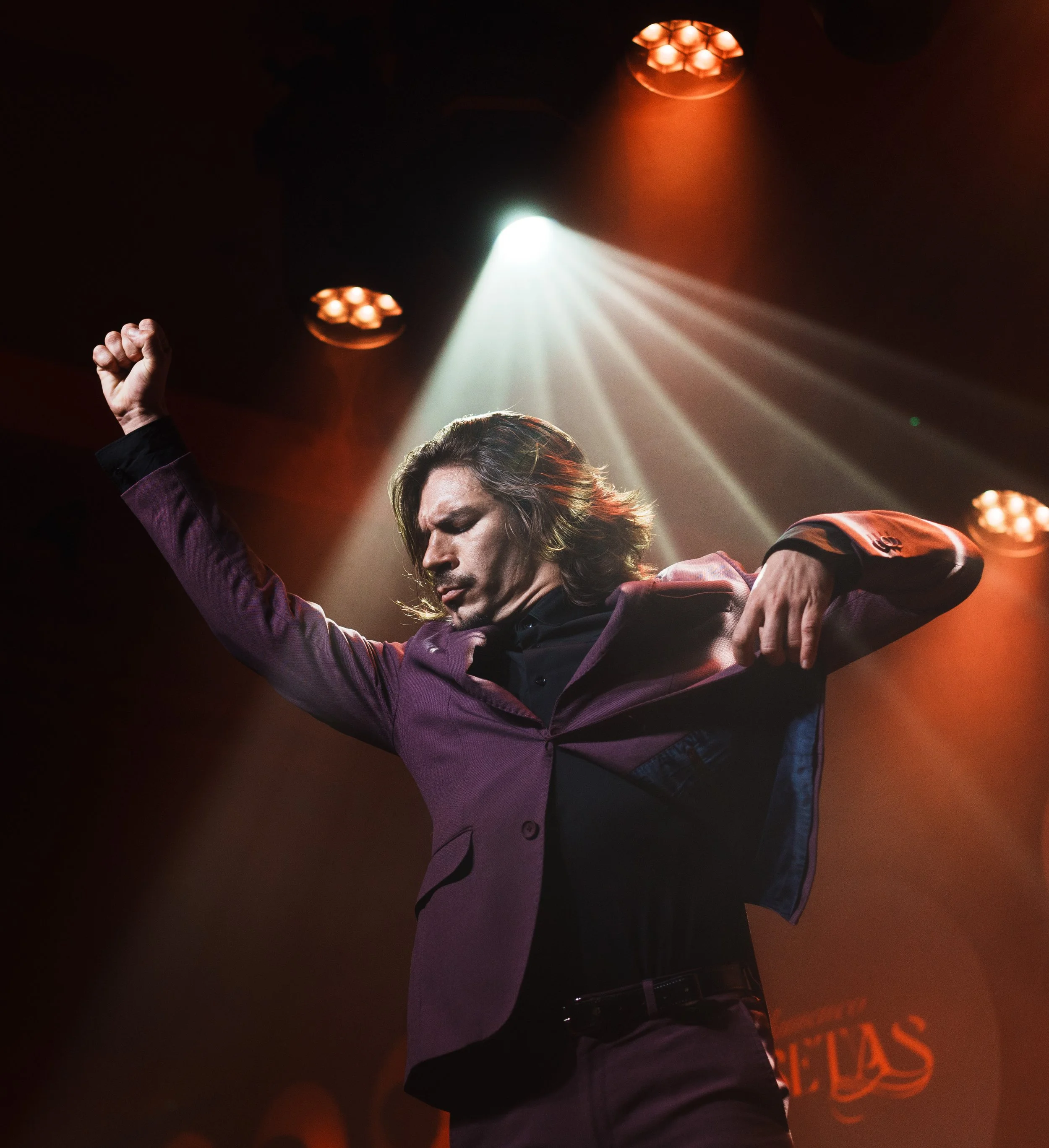 A man with long hair and light skin raising his fist in the air on stage, illuminated by stage lights with orange and white beams.