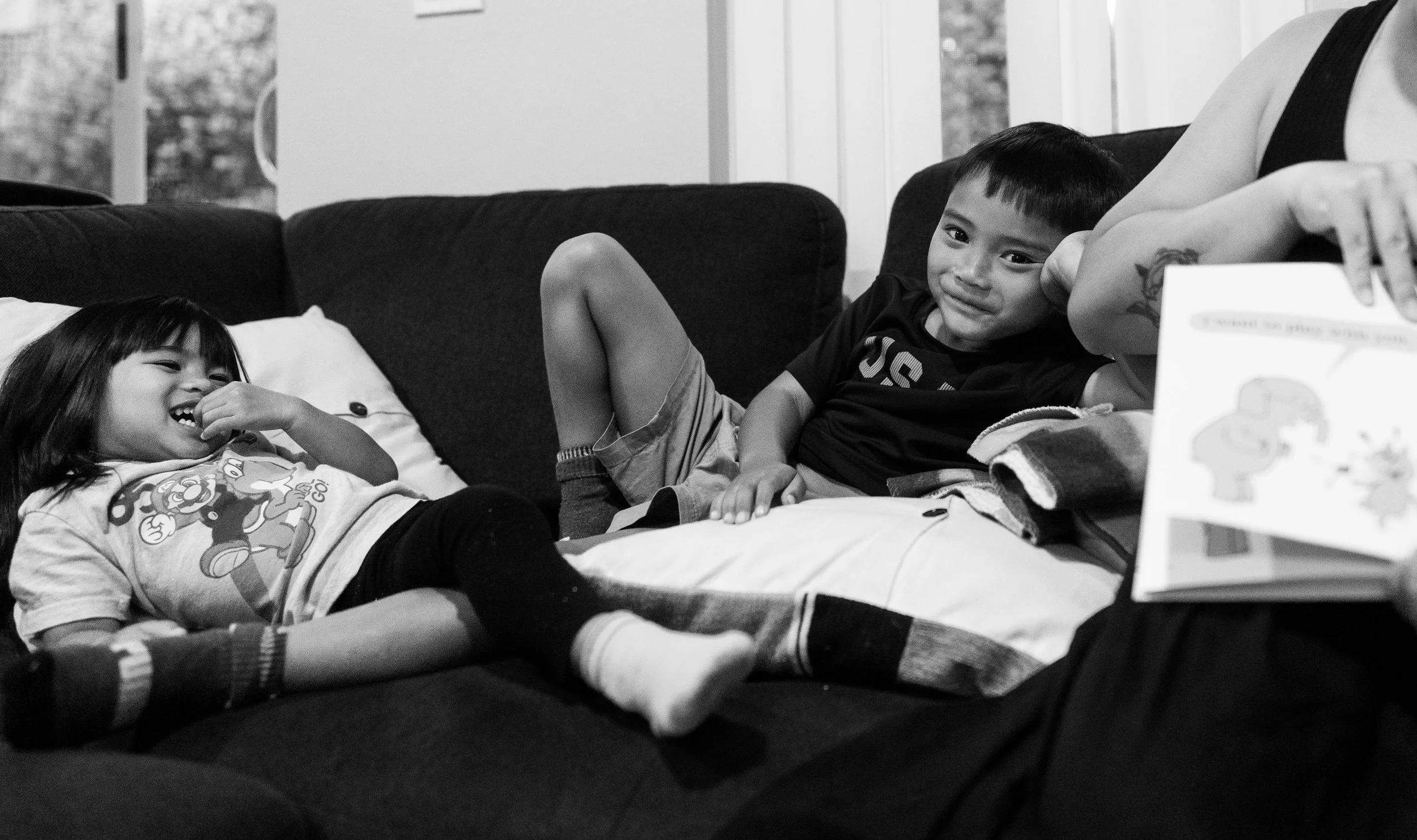 Two children, a girl and a boy, are lying on a sofa, smiling and engaging with each other. The girl has dark hair with bangs and is wearing a cartoon character shirt, while the boy has short hair and is wearing a dark T-shirt. A hand and part of an a