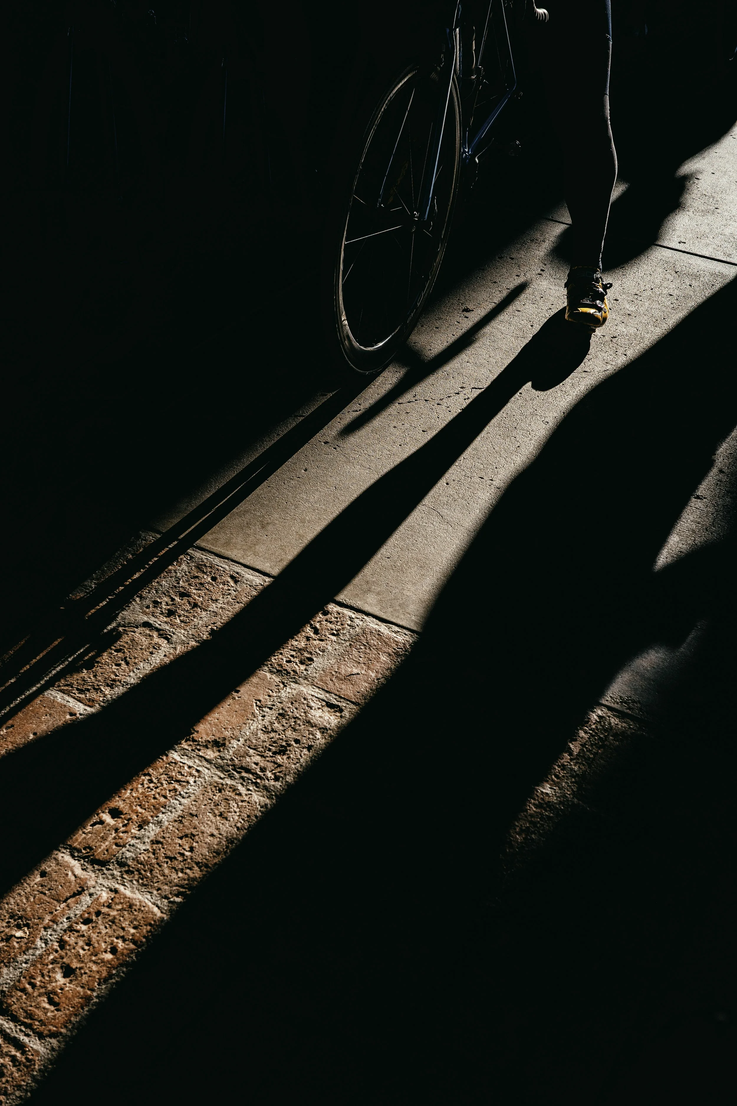 Person walking with a bicycle on a sidewalk, sunlight creating long shadows, dark background.