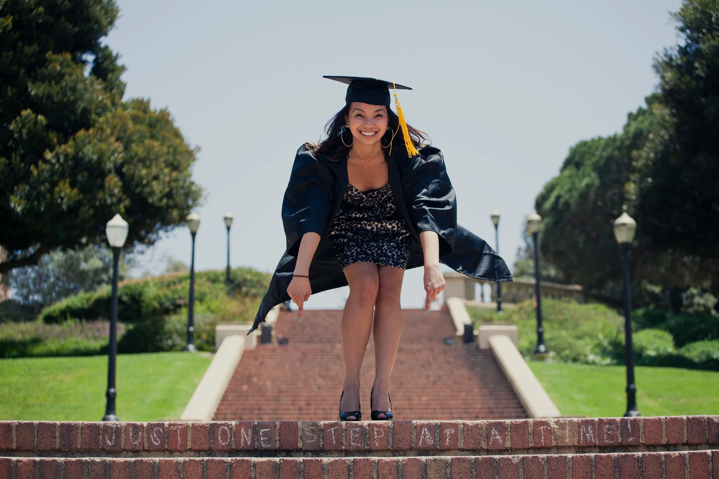 Young woman in a graduation cap and gown posing leaning forward on a brick ledge with the words "POST NO BILLS" partially visible, outdoors in front of a staircase and trees.