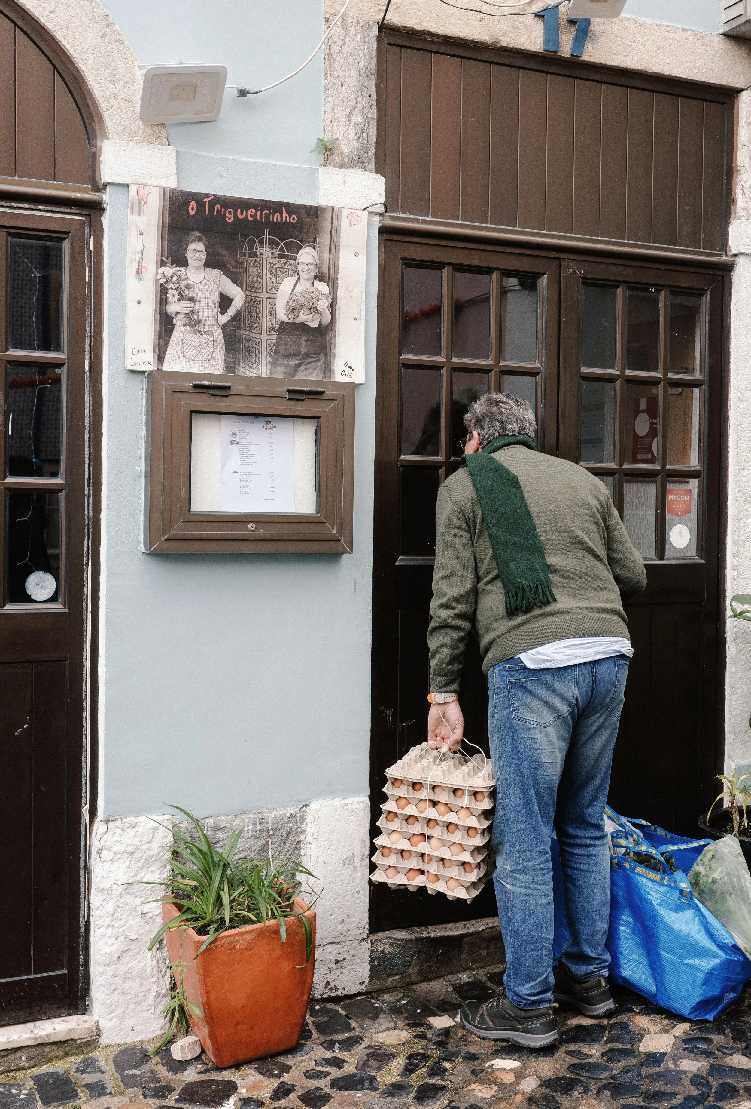 A man with gray hair, glasses, in a green sweater and blue jeans, is holding a carton of eggs and a blue shopping bag, standing outside a building with a black wooden door. There is a large potted plant with green leaves in an orange pot and a black 