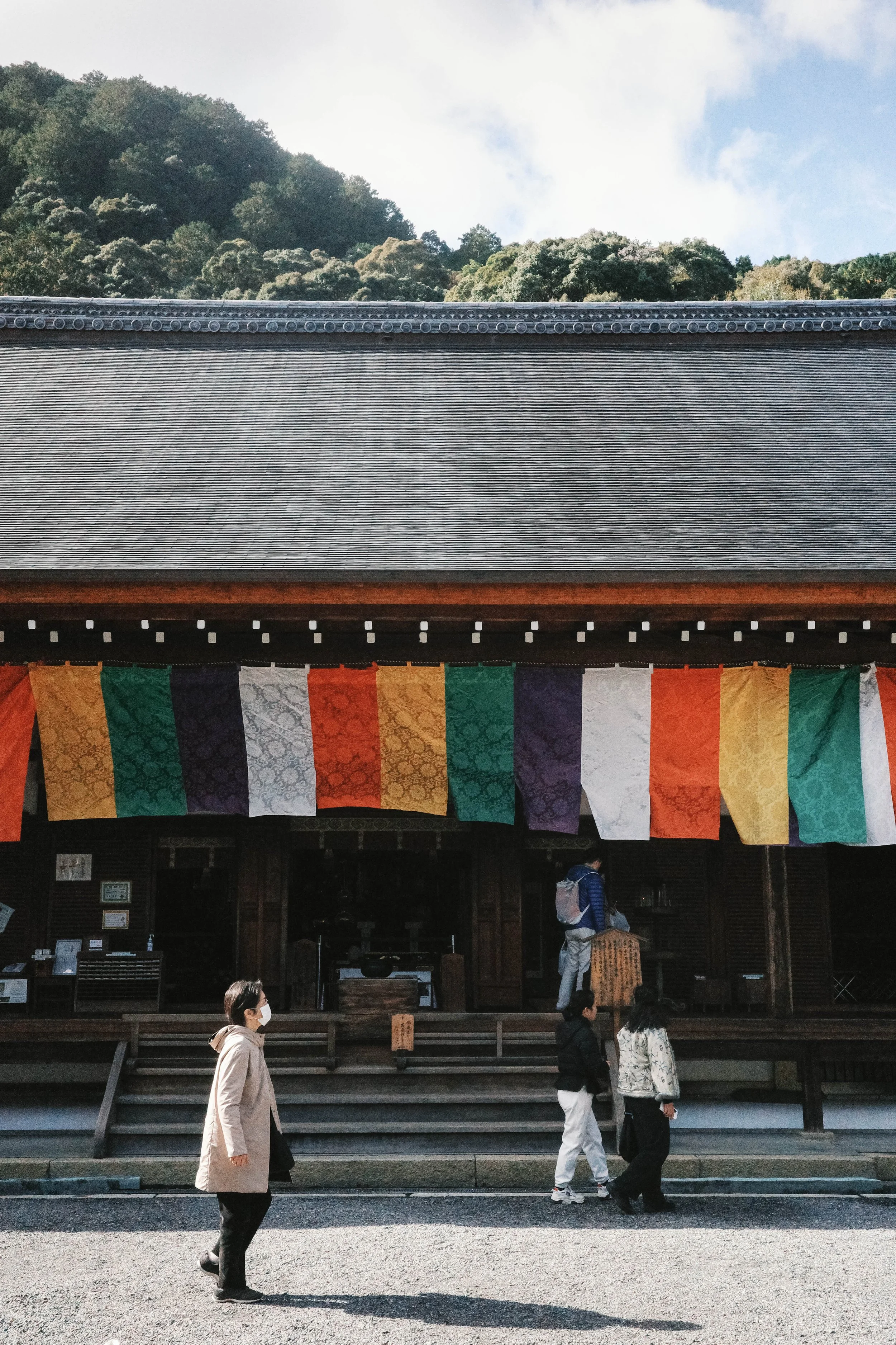 People walking in front of a Buddhist temple with colorful prayer flags and a traditional tiled roof.