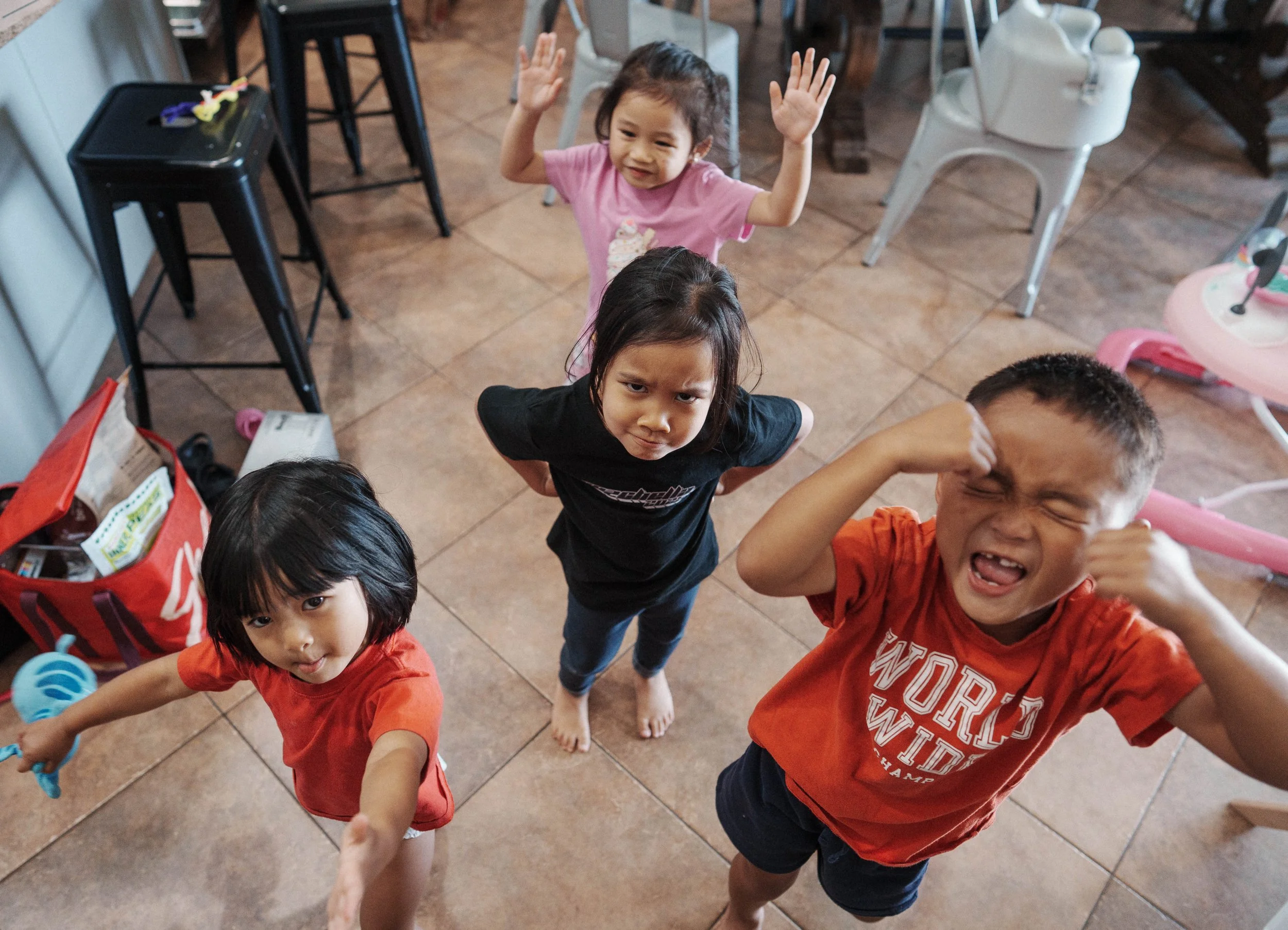 Group of five children standing on a tiled floor, looking up at the camera, some with hands raised or making expressions, with chairs and a high chair in the background.