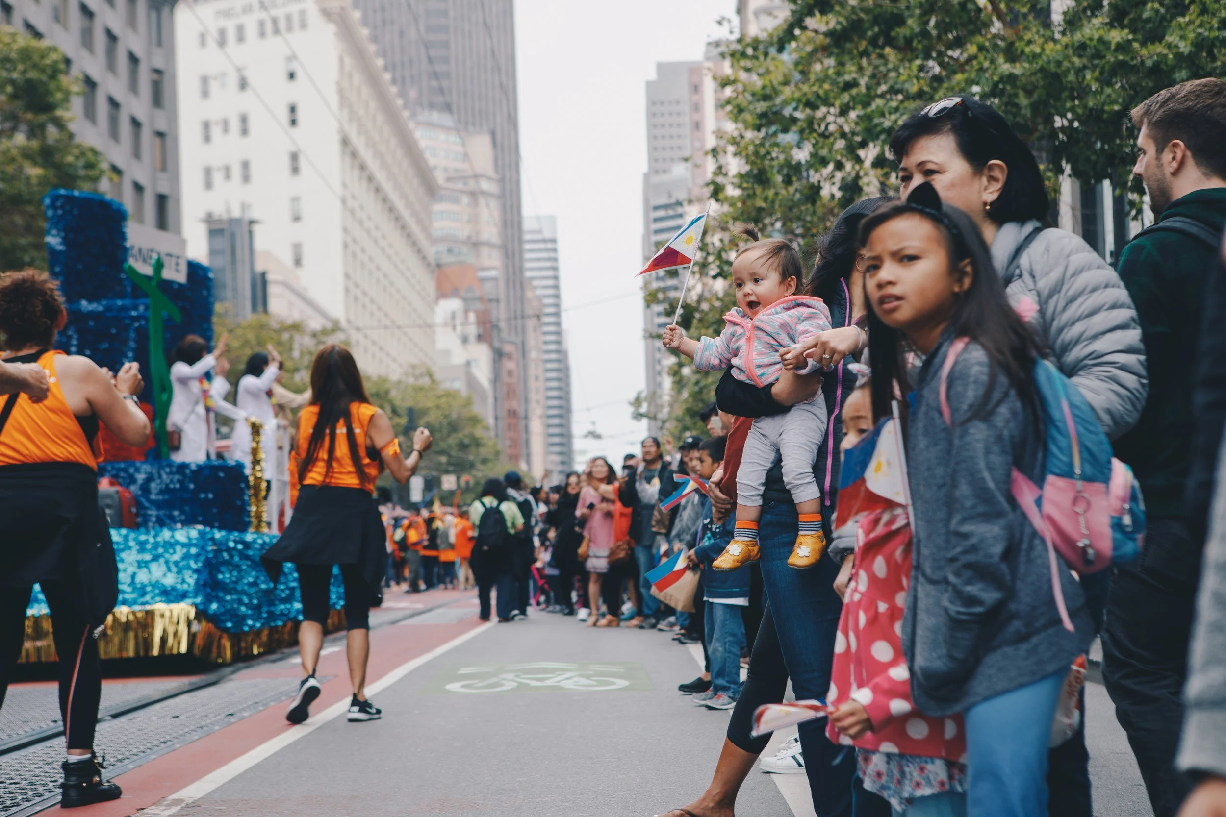 People participating in an outdoor parade or event on a city street, with tall buildings in the background. Children and adults are watching or taking part, some holding small flags, and decorated floats and decorations are visible.