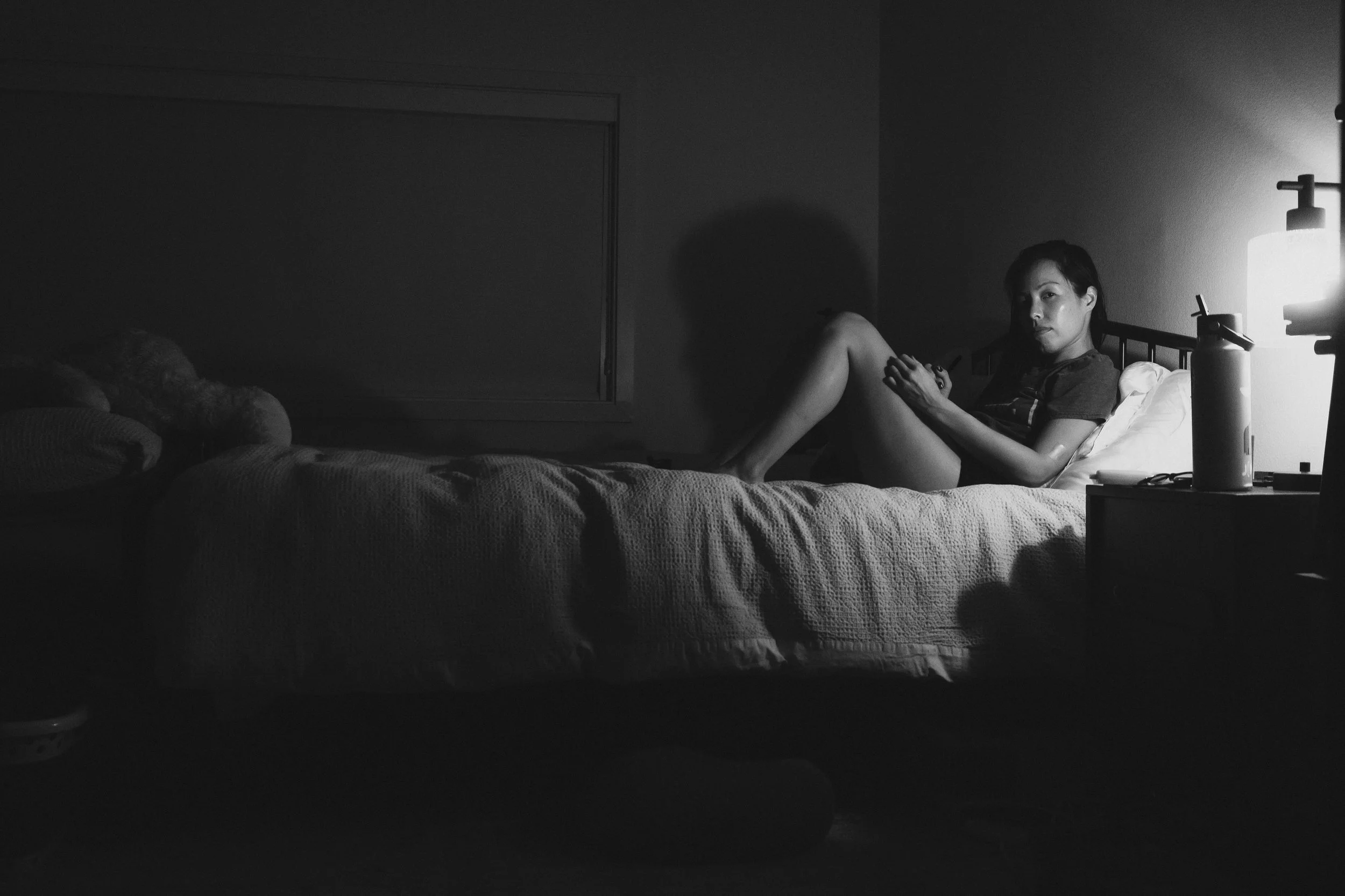 A woman sitting on a bed in a dimly lit room, illuminated by a bedside lamp on the nightstand, with pillows and a blanket on the bed.