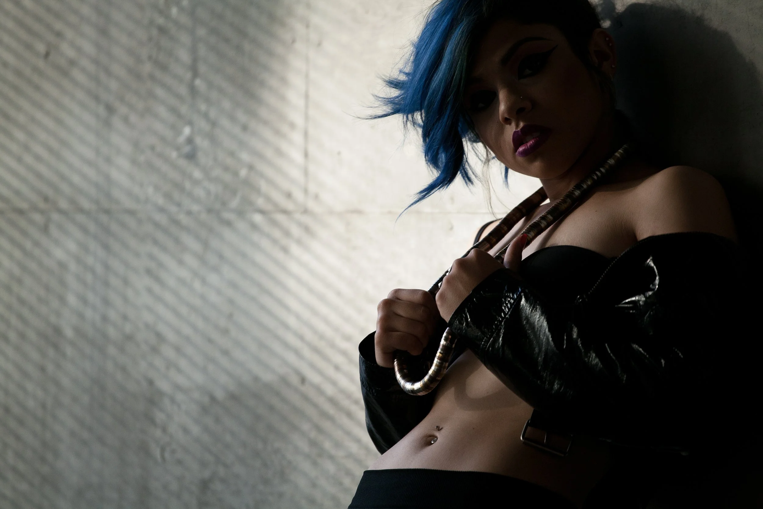 A woman with blue hair styled in a messy bob, wearing black makeup and lipstick, with multiple earrings and piercings, holding a decorative cane or staff, dressed in a black leather jacket and exposing her midriff against a textured light gray wall.