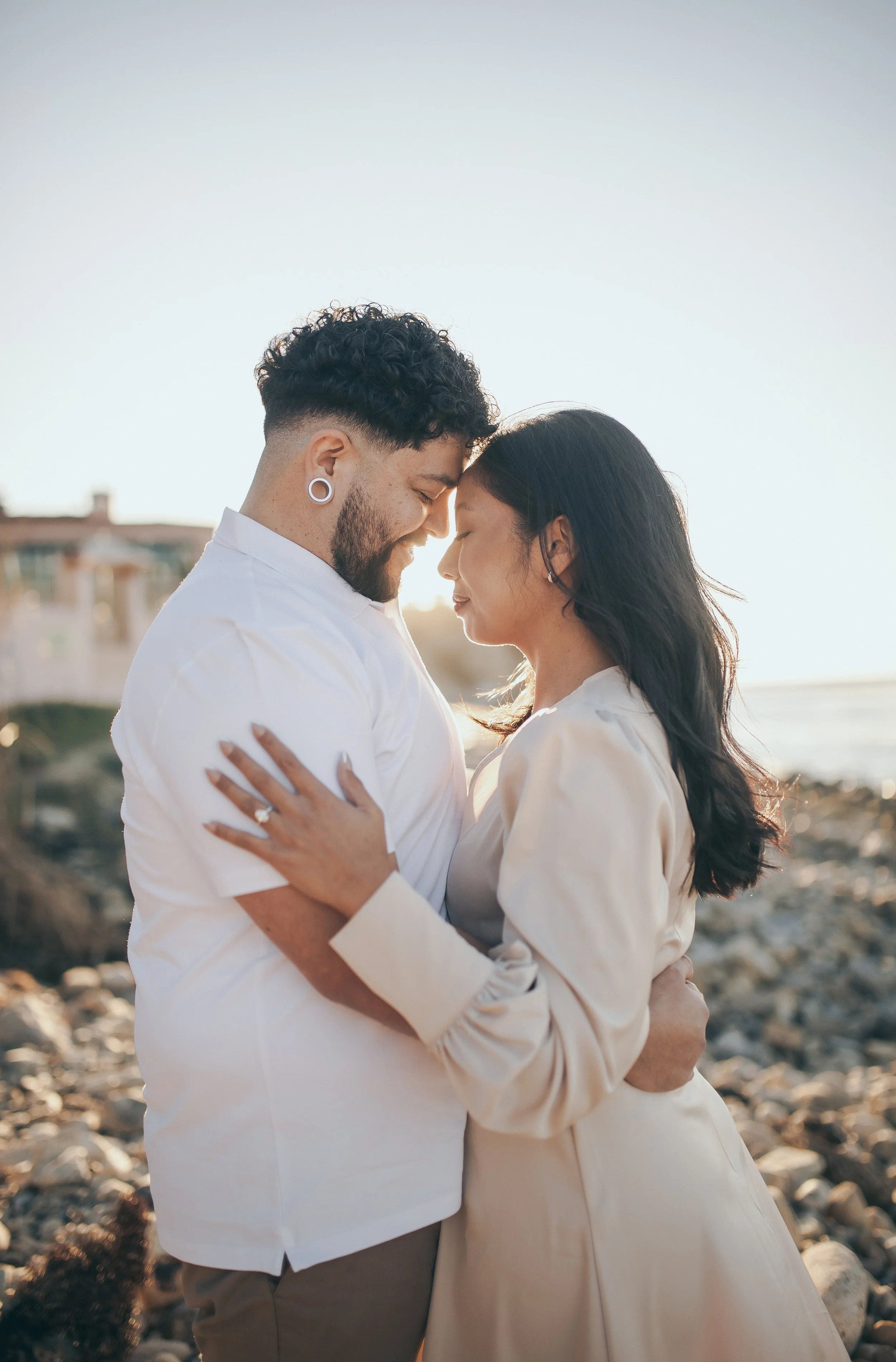 A couple standing close with their foreheads touching on a rocky beach at sunset, embracing with eyes closed in a romantic moment.