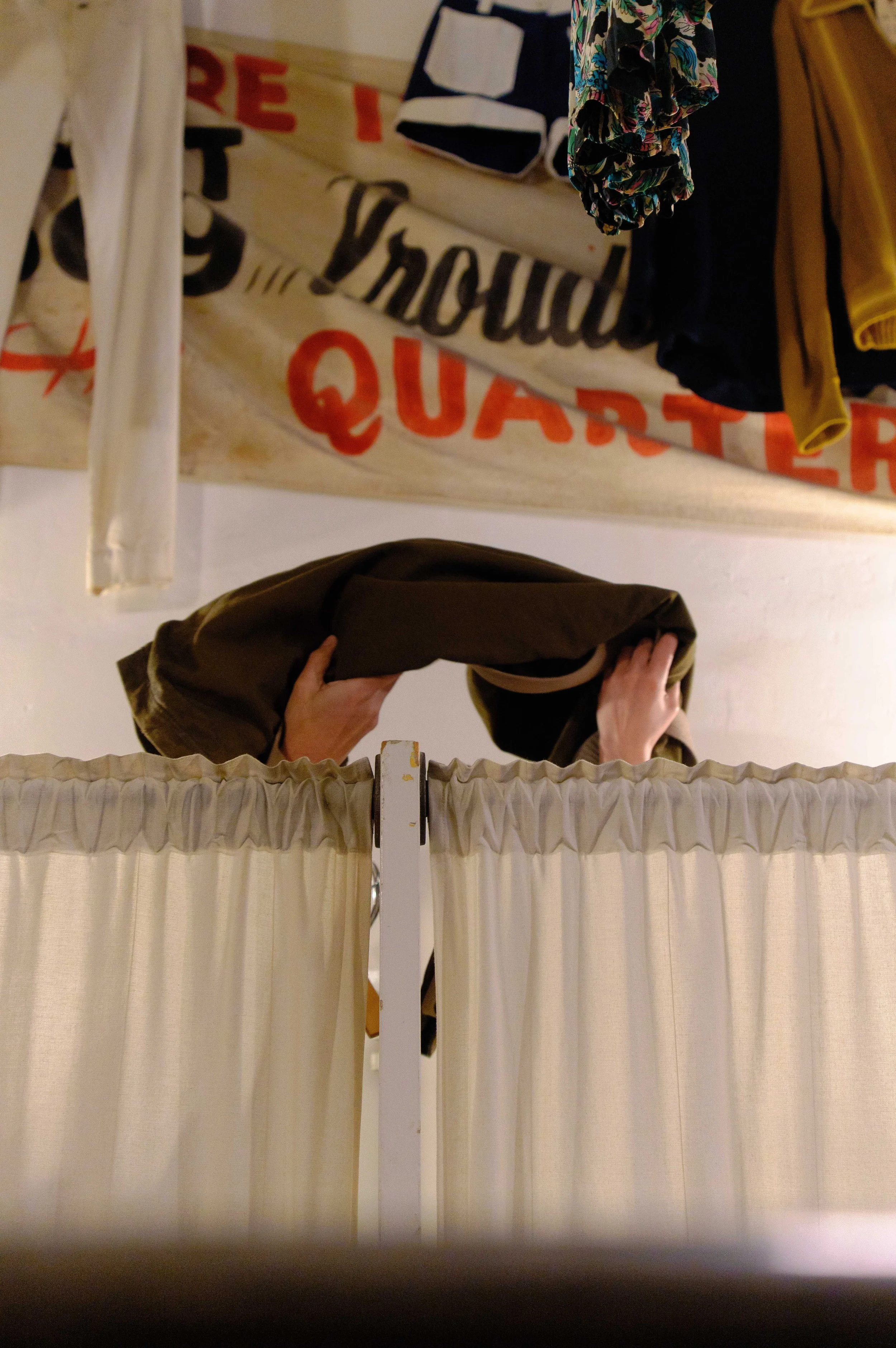 Mirror reflection showing hanging clothing items behind a beige curtain. The person taking the photo is partially visible, holding a dark-colored bag.