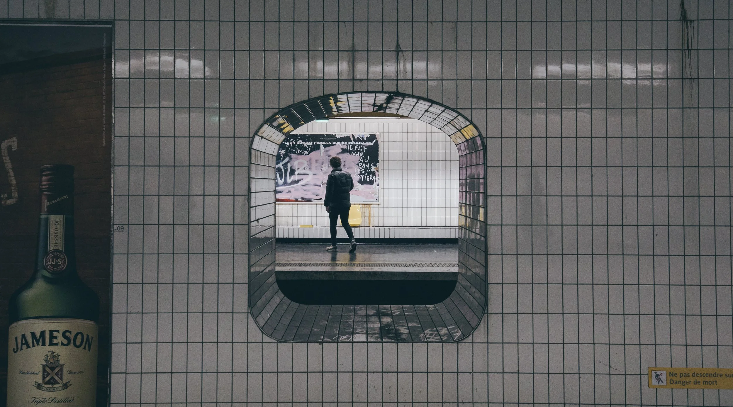 A person standing in front of a large advertising poster in a tiled subway station. There is a bottle of Jameson Irish whiskey on the left and a yellow warning sign on the bottom right corner.