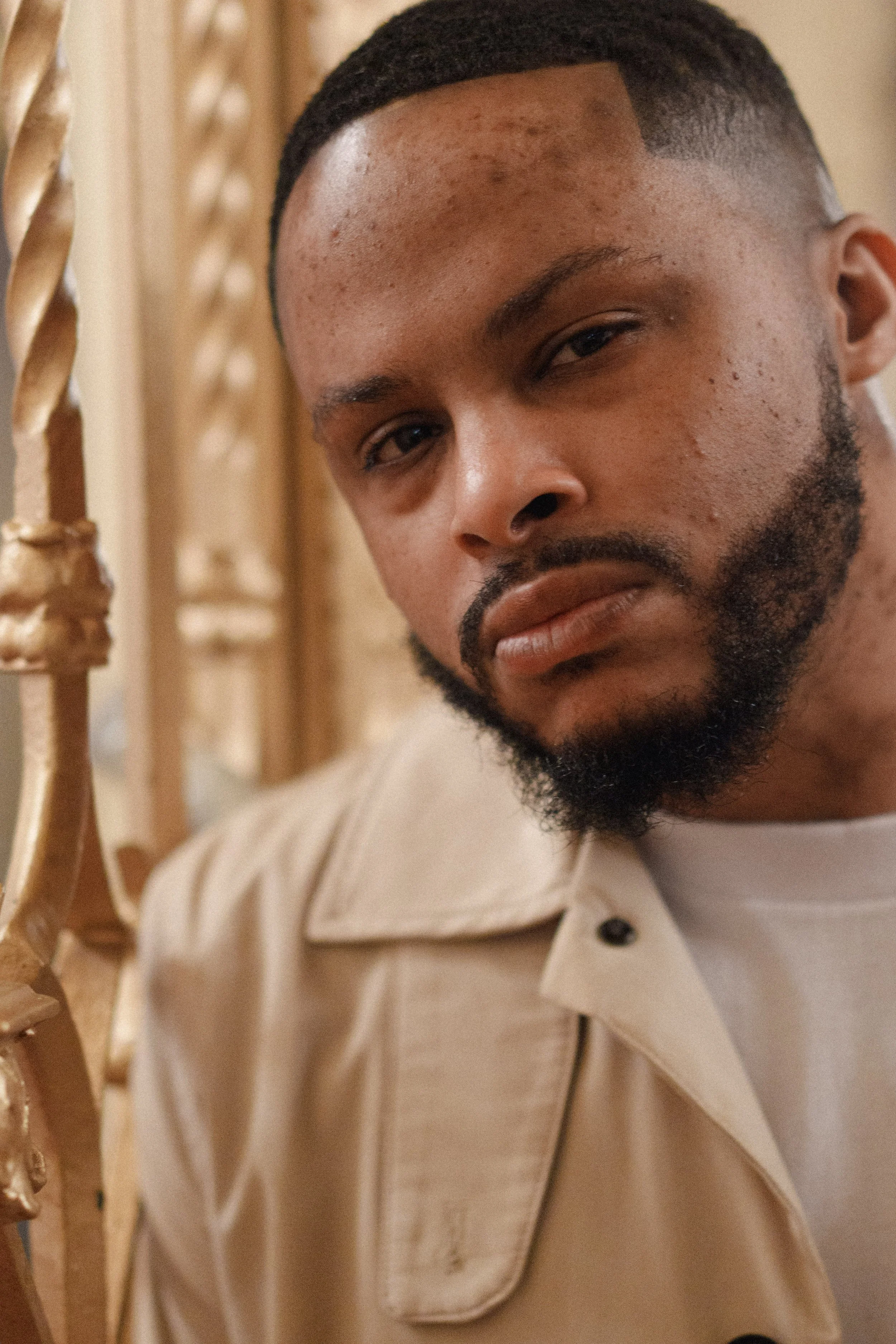 Close-up of a man with a beard and short hair, wearing a beige jacket, standing next to a wooden railing or decorative structure. He has a serious expression, and the background is blurred.