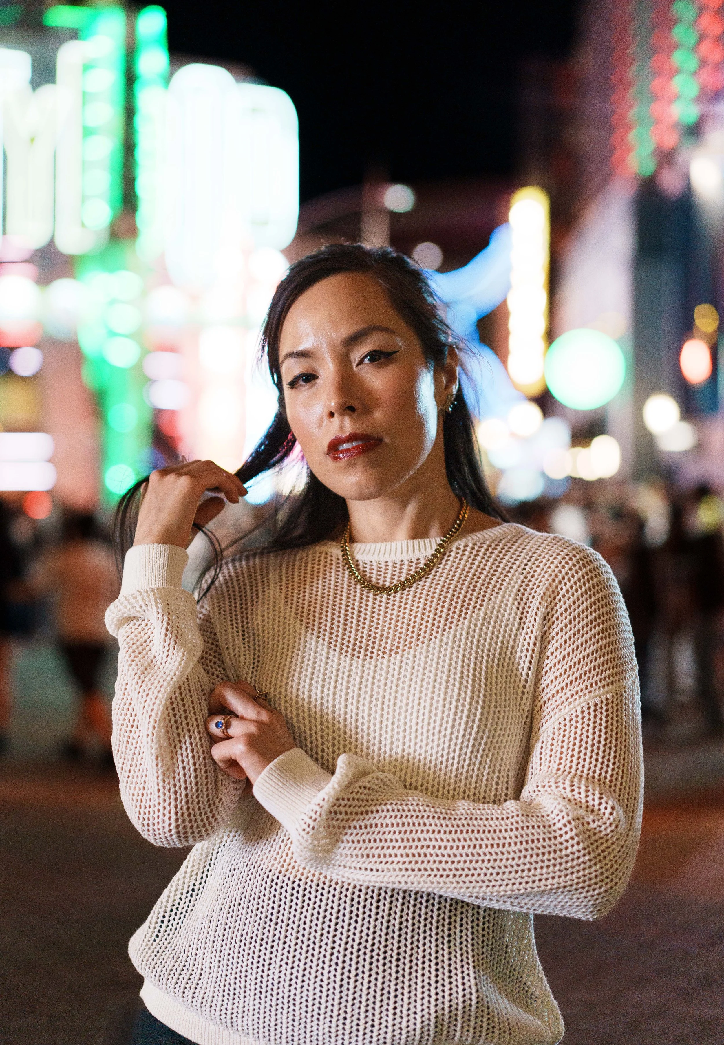 A woman with black hair and makeup standing in front of colorful neon lights at night, wearing a loose knit beige sweater, gold chain necklace, and small earrings, with a confident expression.