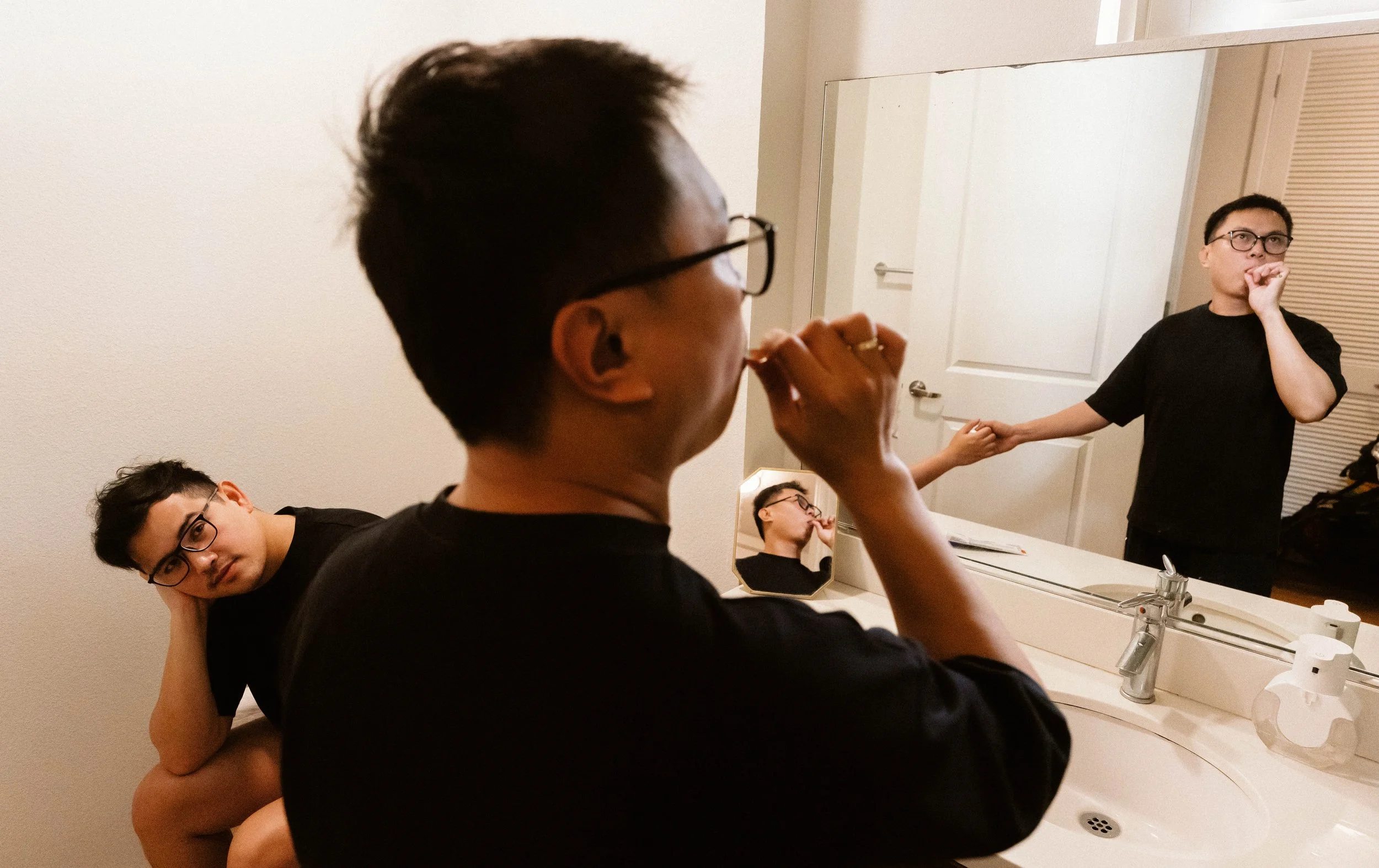 A woman standing at a bathroom mirror, wearing glasses and a black shirt, with her hand near her face. Two men are also in the scene, one sitting and the other with his head tilted, both wearing glasses and black shirts, with one resting his arm on t