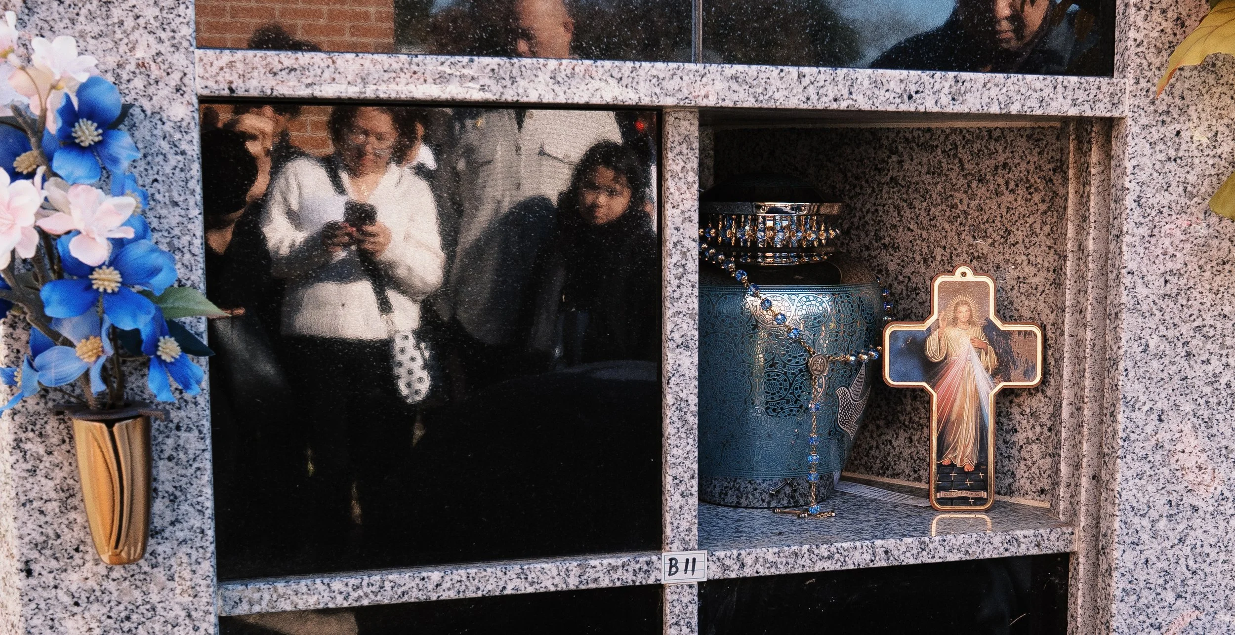 A granite memorial wall with a photo of a woman smiling and looking at a phone, a blue vase with pink and blue artificial flowers, a crucifix with Jesus, and various religious items including a rosary and a small statue of Jesus surrounded by a cross