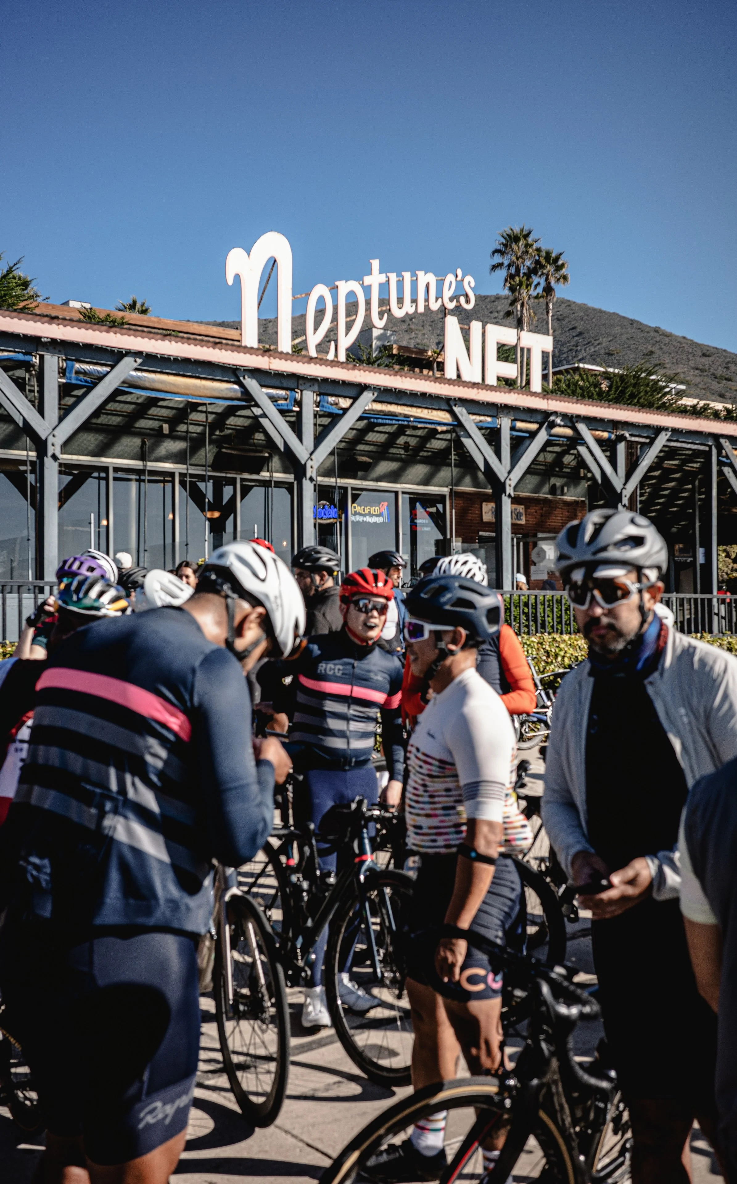 Group of cyclists in cycling gear and helmets gathered outside a building with a sign that says 'Neptune's Net' and palm trees in the background.
