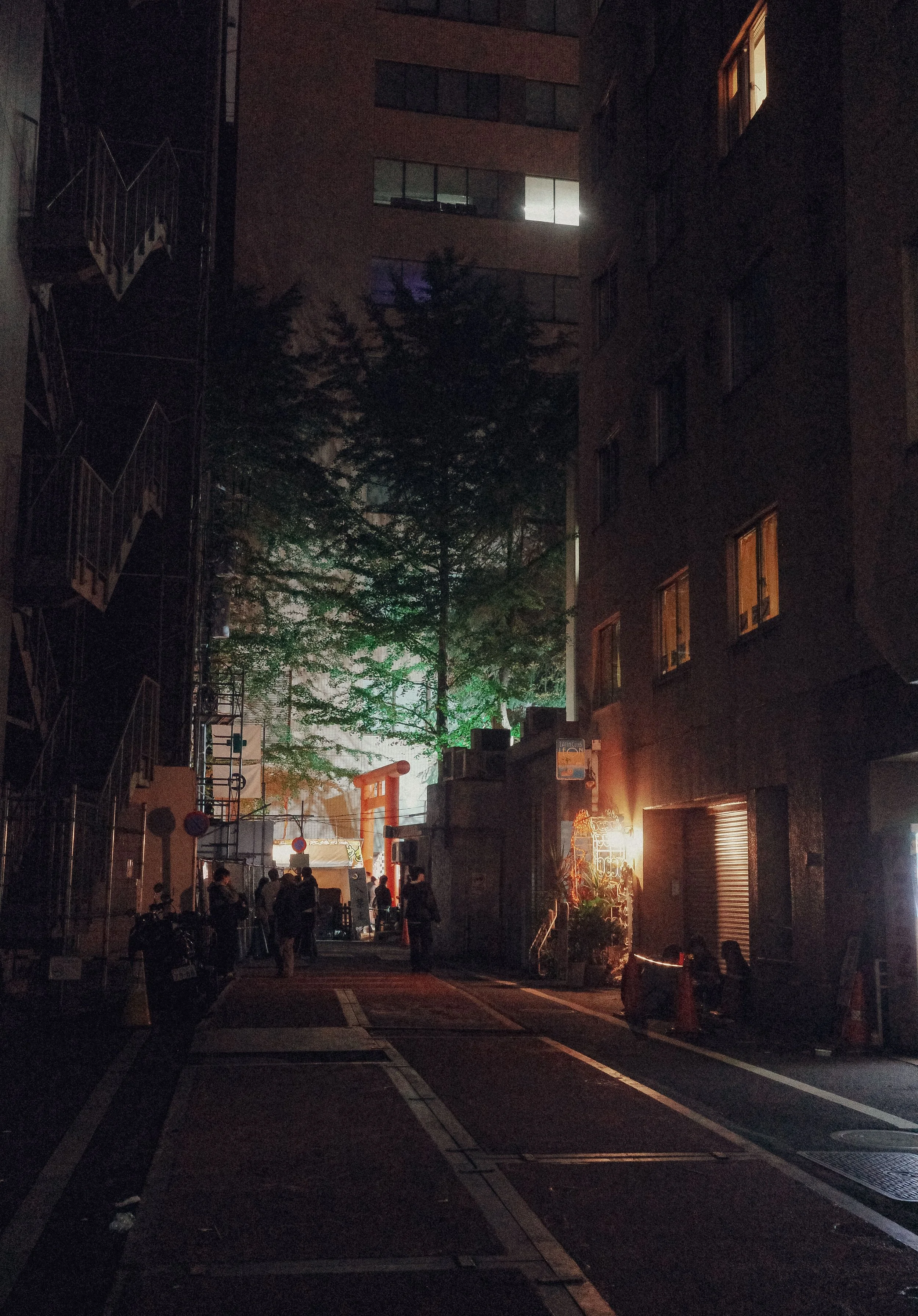Night scene of a city street with some people gathered near a lit storefront. Tall buildings with lit windows line the street. A tree and some street signs are visible, and there's a lit torii gate in the background.