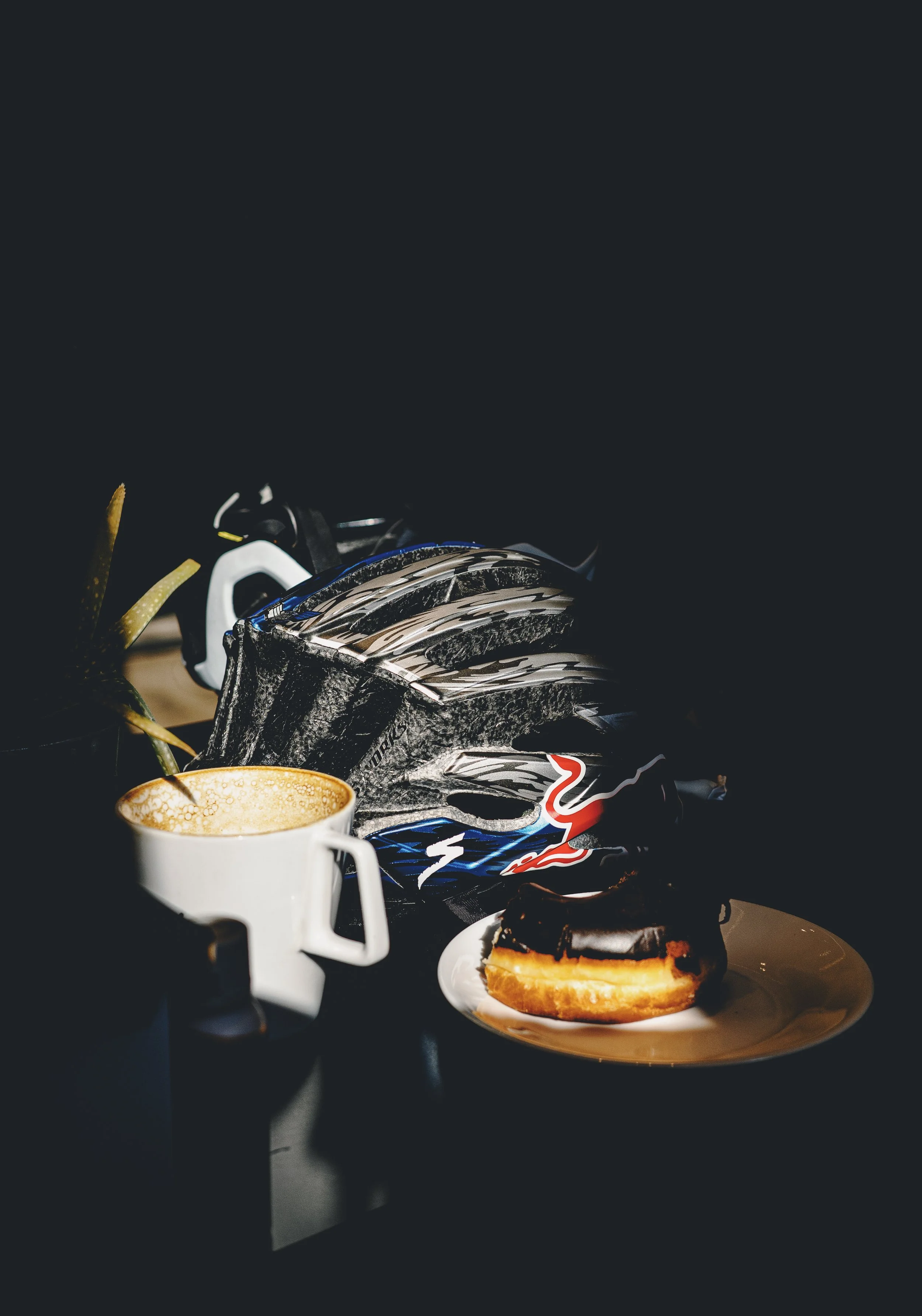 A coffee mug with foam art, a chocolate cake, and a plate with a cookie sandwich in front of a black and white motocross helmet and a black bicycle helmet on a dark surface against a dark background.